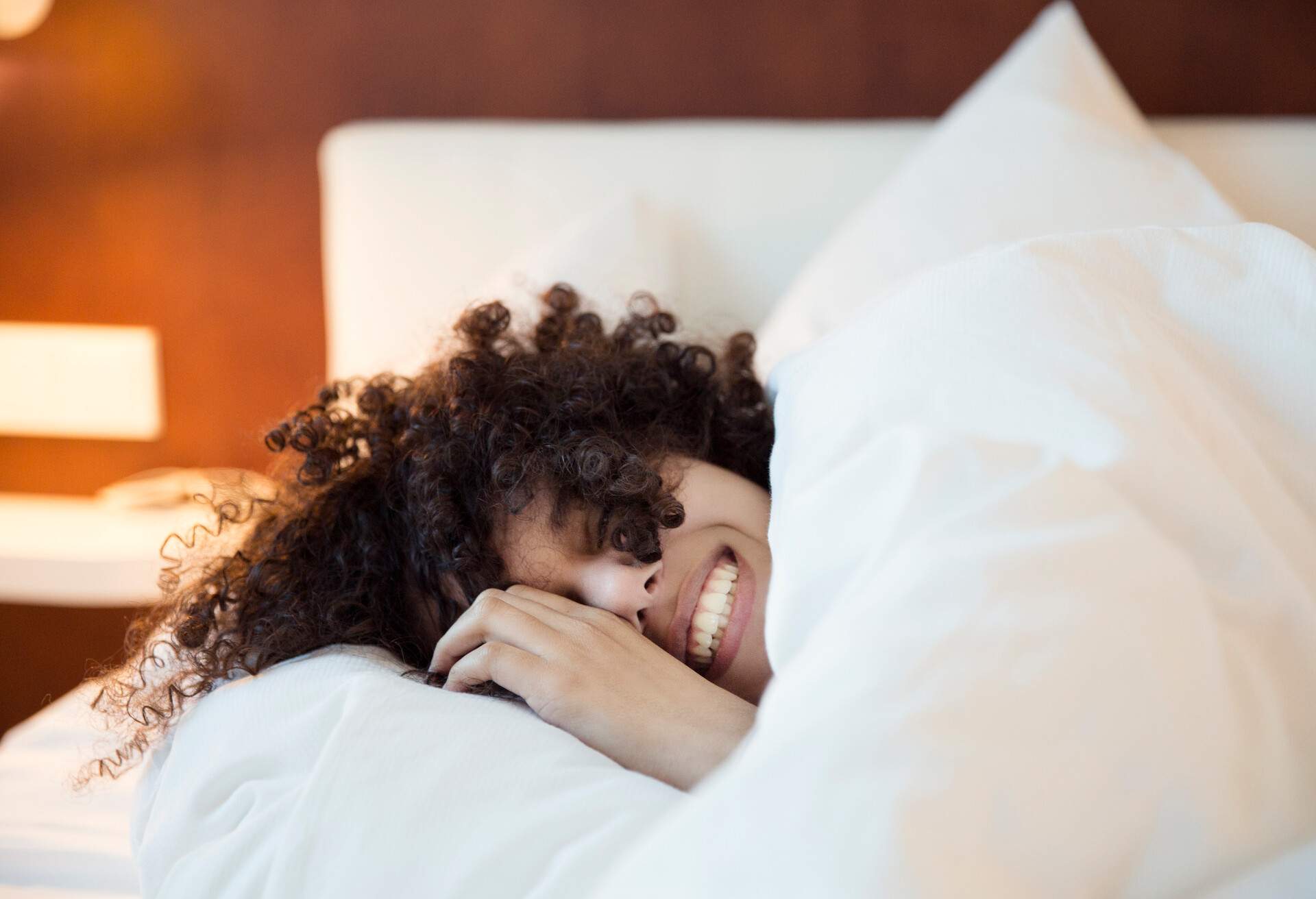 A curly-haired woman smiles as she snuggles under a blanket.