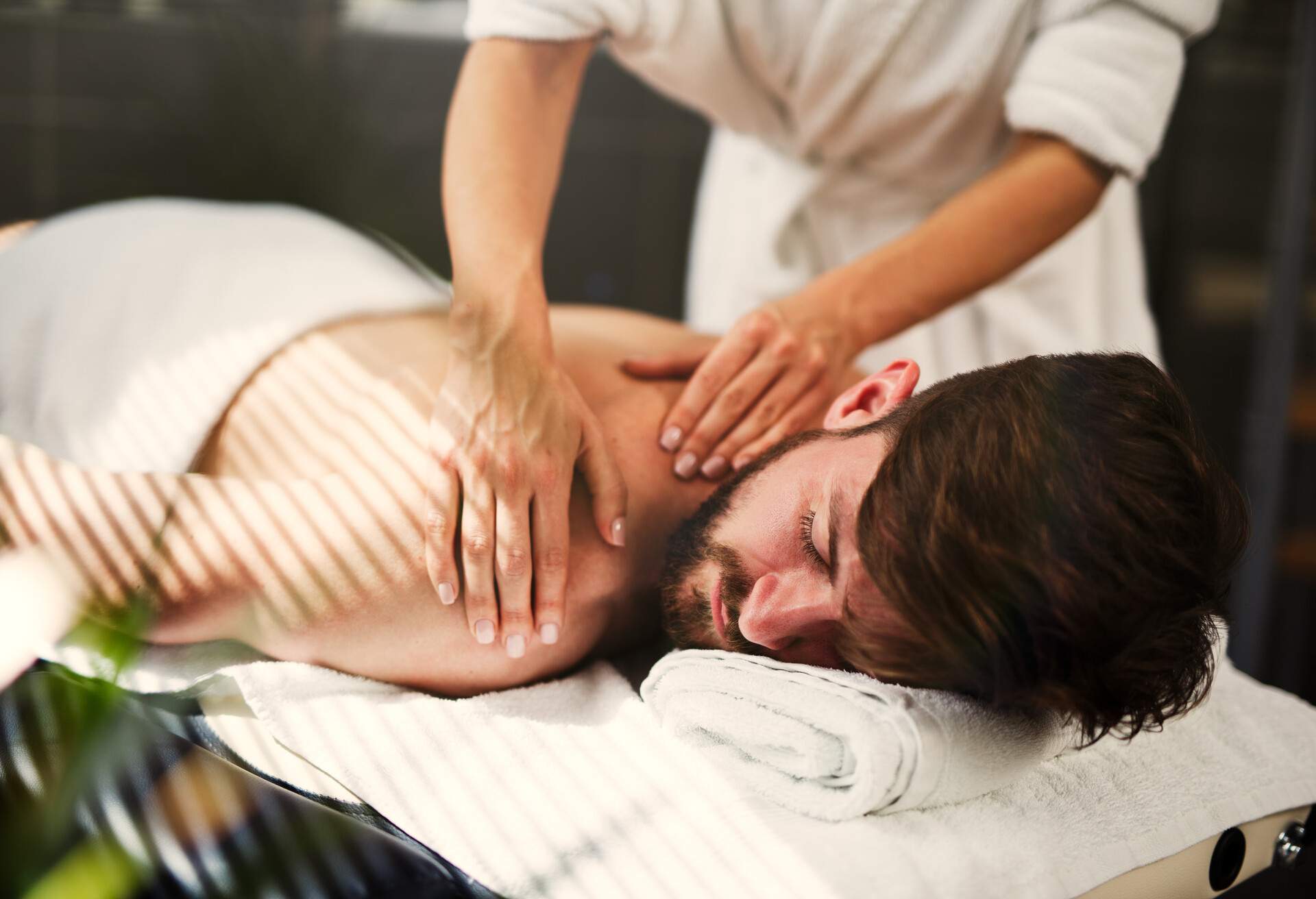 A bearded man lies on a massage table as she receives a back massage.