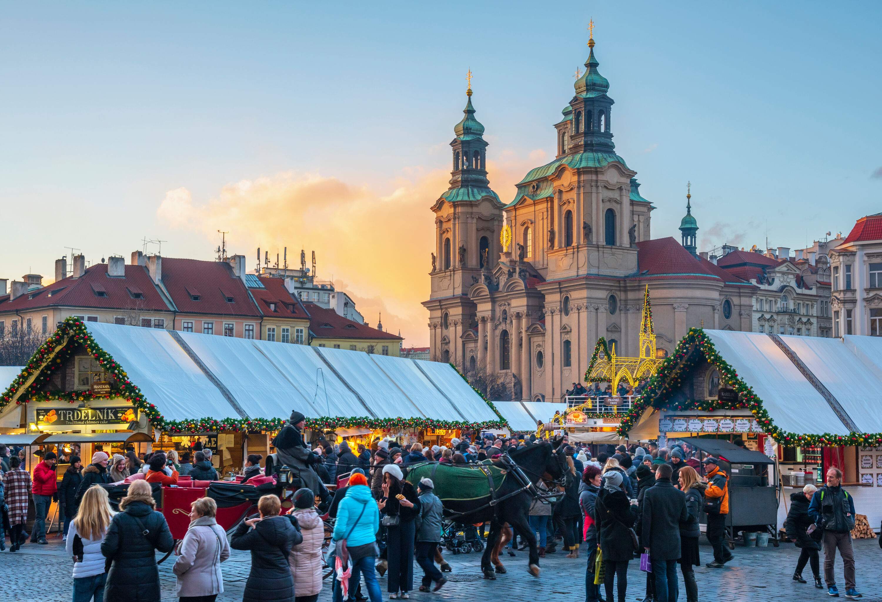 A crowd of people in the Christmas market with stalls decorated with Christmas ornaments, a horse with a red carriage, and the Baroque Church of Saint Nicholas in the background.