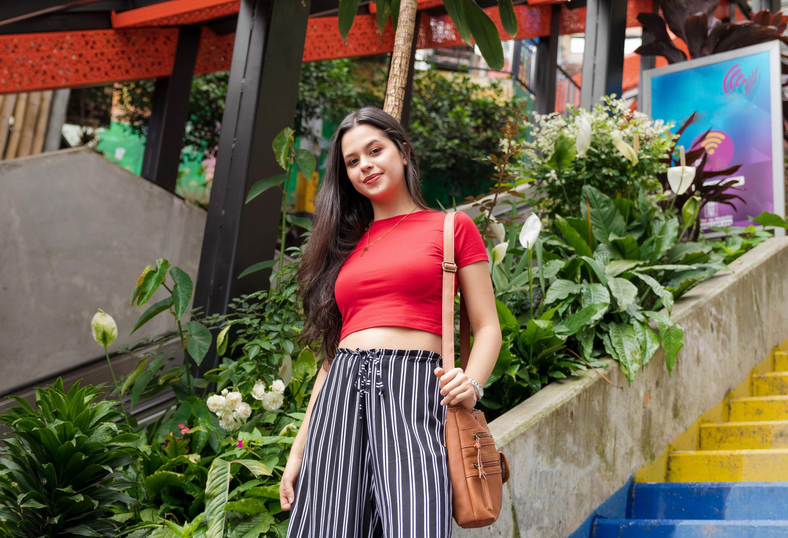 A young Latina, her long hair flowing, wearing a red shirt with a radiant smile, gracefully descending a stair adorned with vibrant blooming plants.