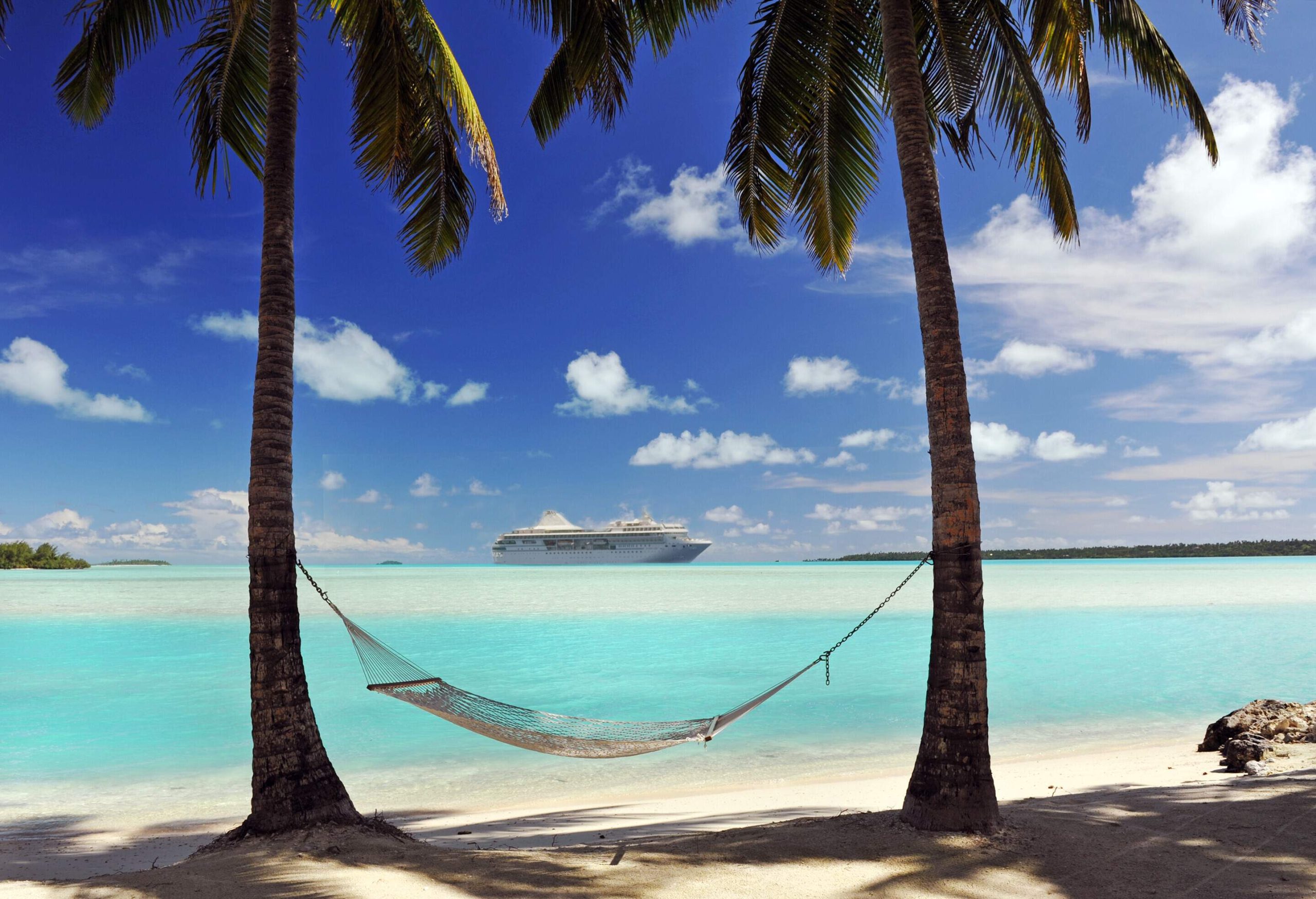 A hammock tied on two palm trees with distant views of a cruise ship in the ocean.