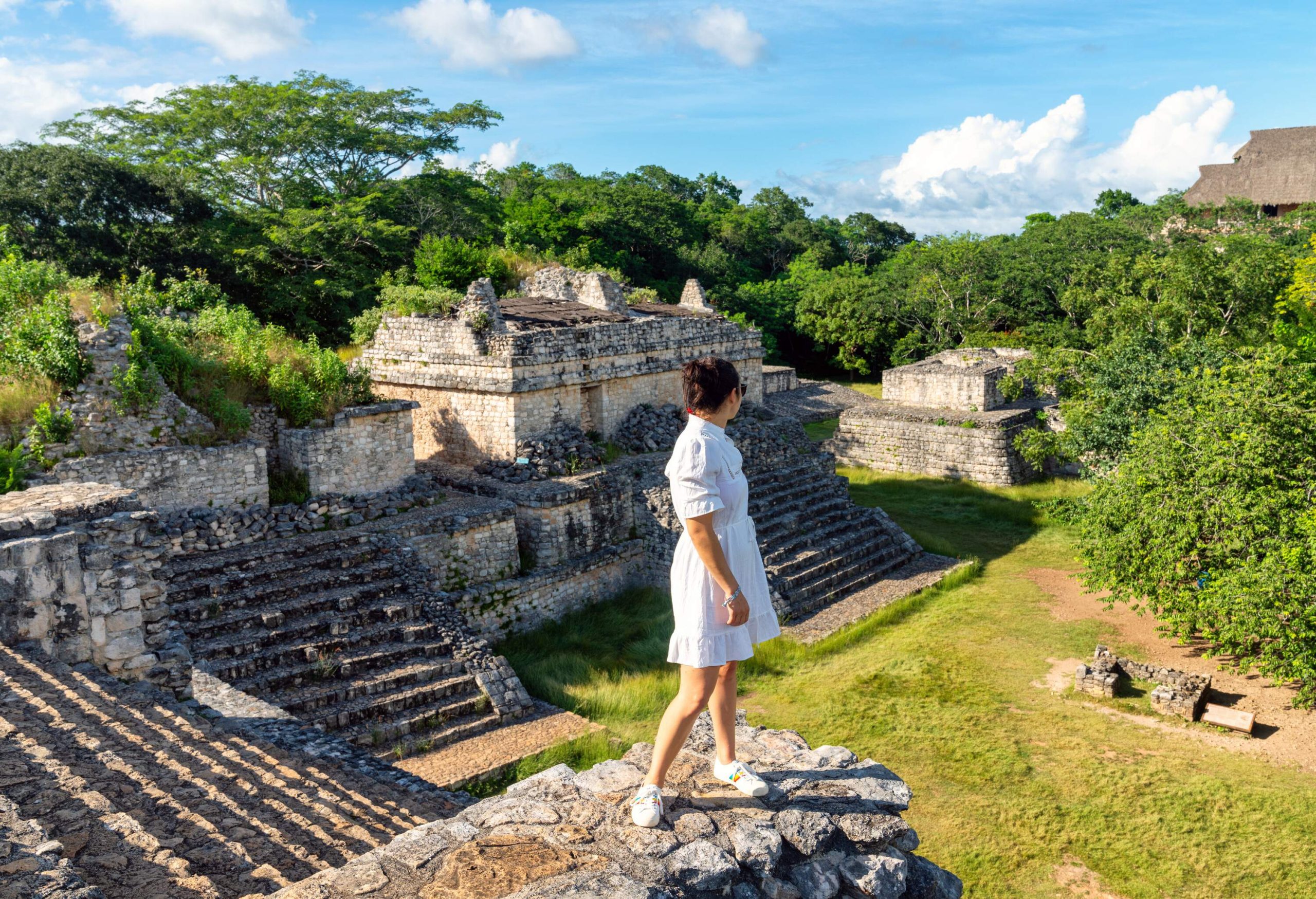 A woman in a white dress stands at the edge of a stone platform admiring the views of Mayan ruins.