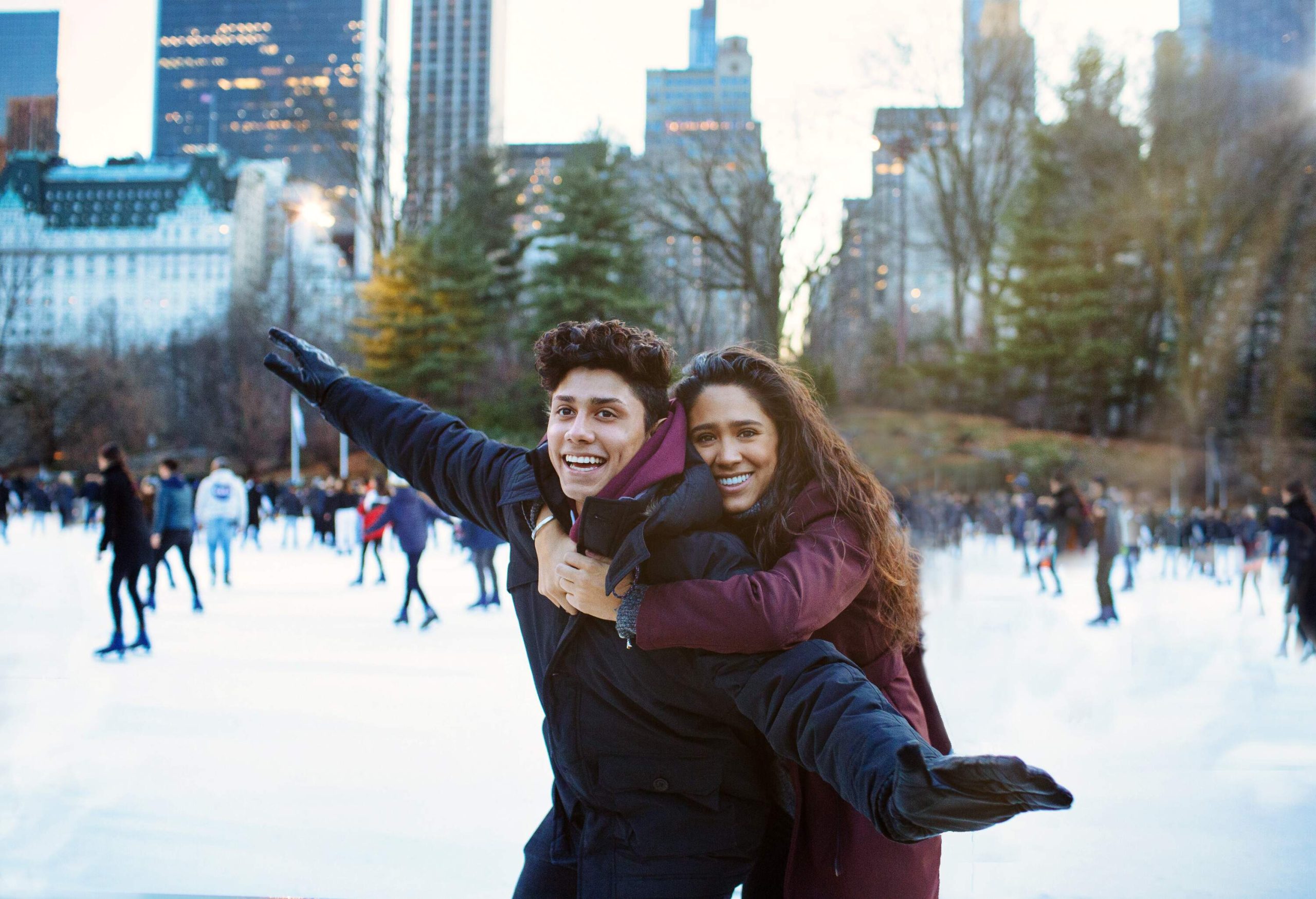 A young lady hugs her man from the back while enjoying skating on the ice outdoors.