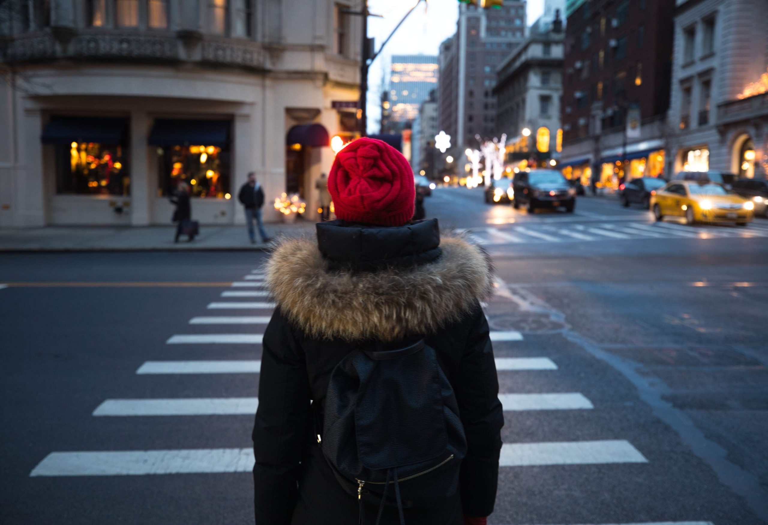 A person in a red bonnet, black fur coat, and bag stands in front of a crosswalk.