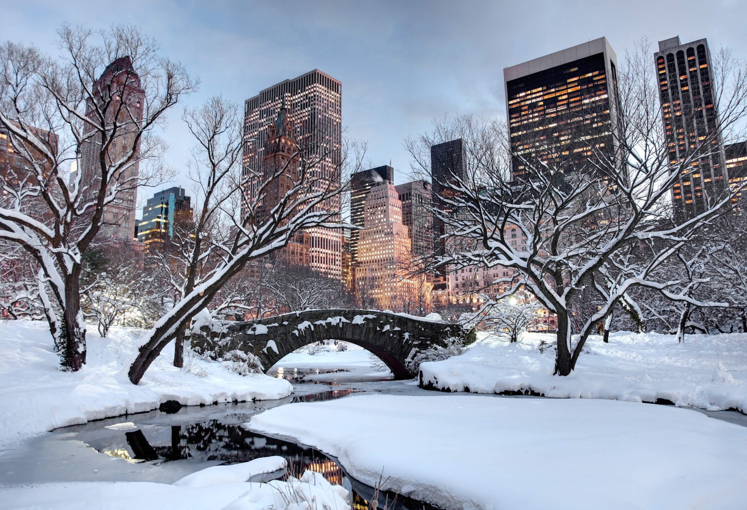 A stone arch bridge across a pond in a snow-covered park with bare trees and views of a city skyline in the background.