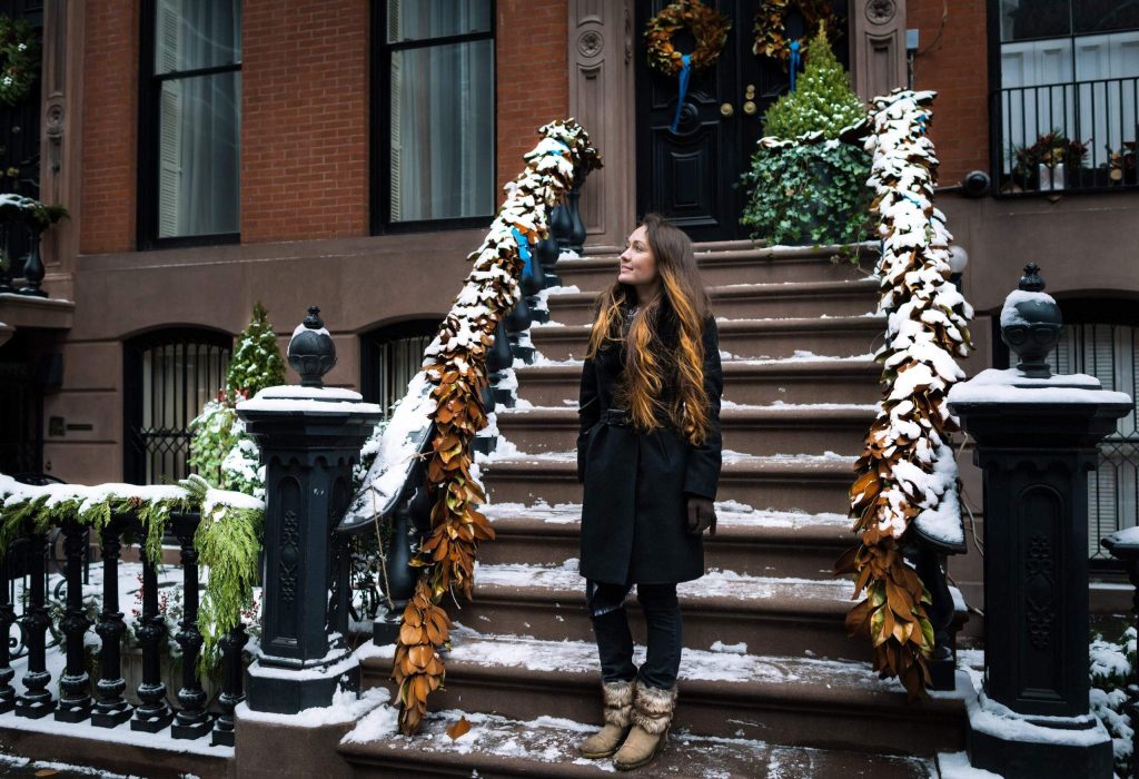 A woman with long brown hair stands on a stoop on the streets of Brooklyn.