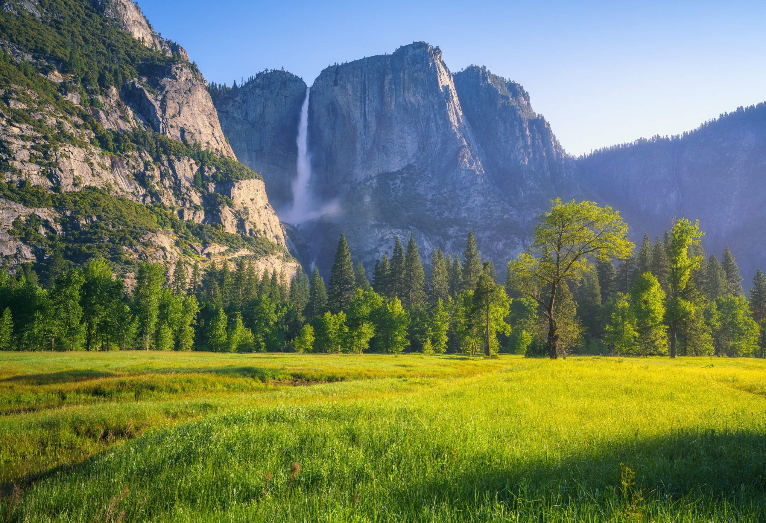 A verdant valley surrounded by tall green trees beneath the ragged mountain range with a waterfall.