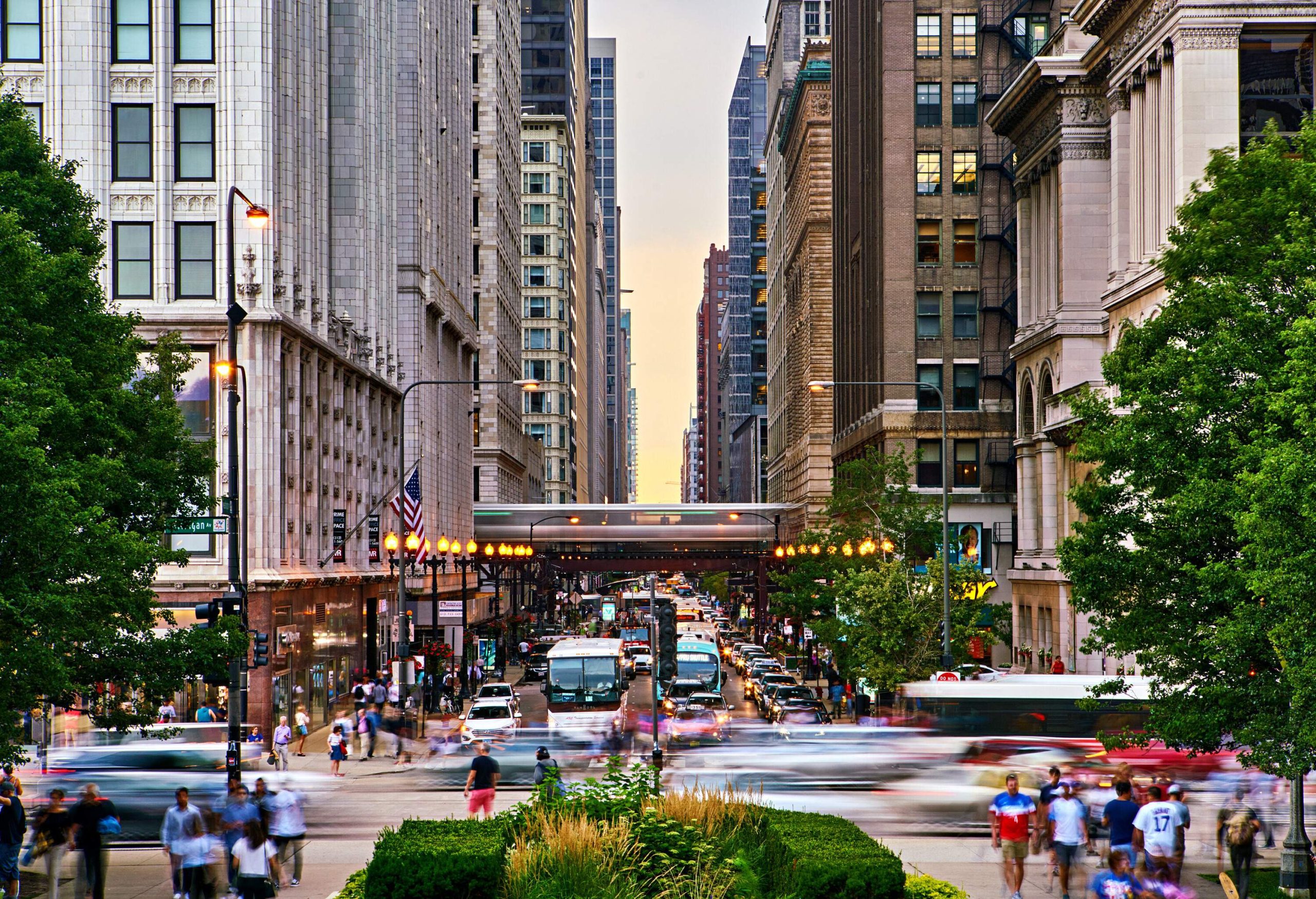 A busy intersection with passing cars and pedestrians surrounded by lush trees and tall city buildings.