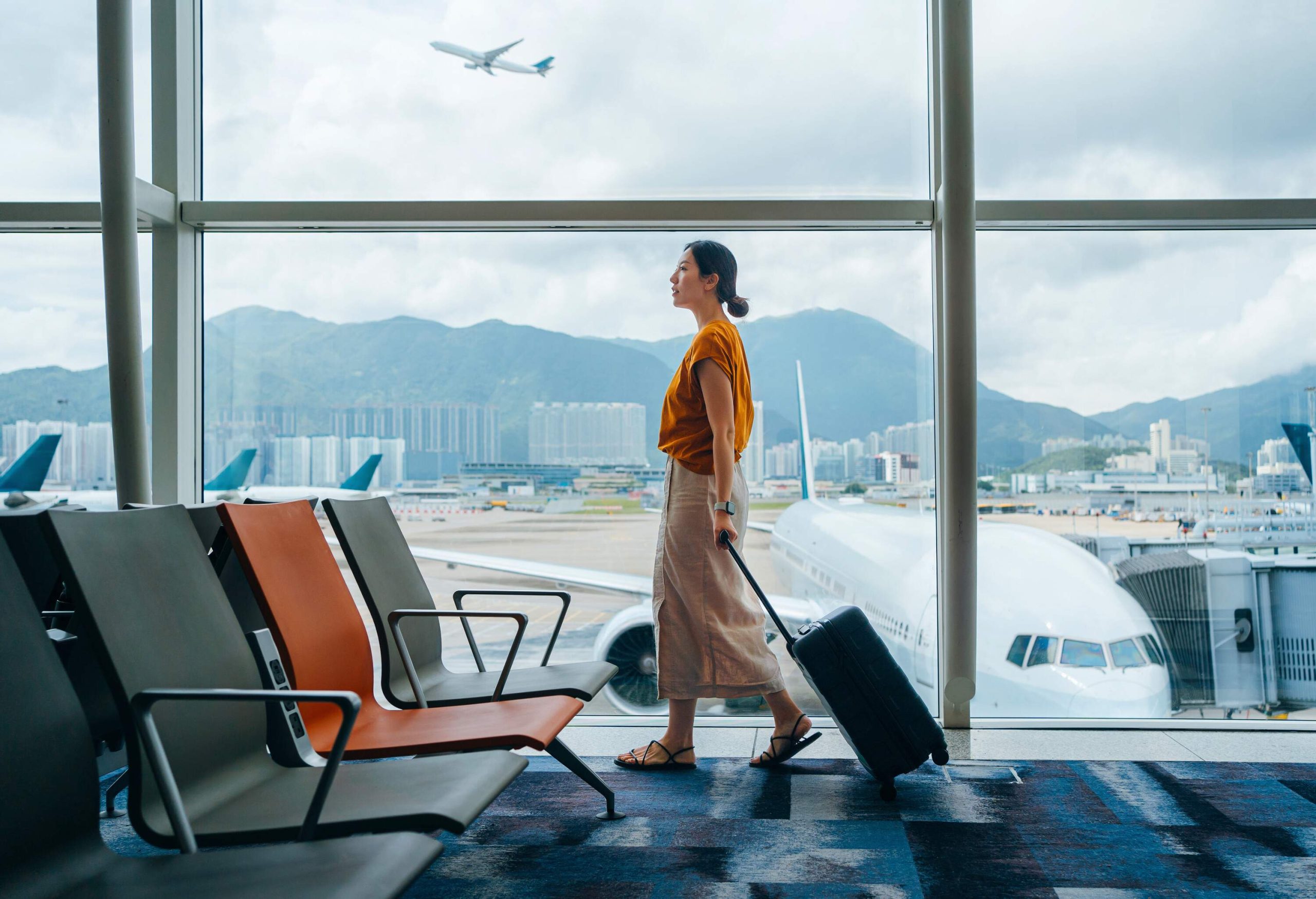 Asian woman carrying suitcase, walking by the window at airport terminal.