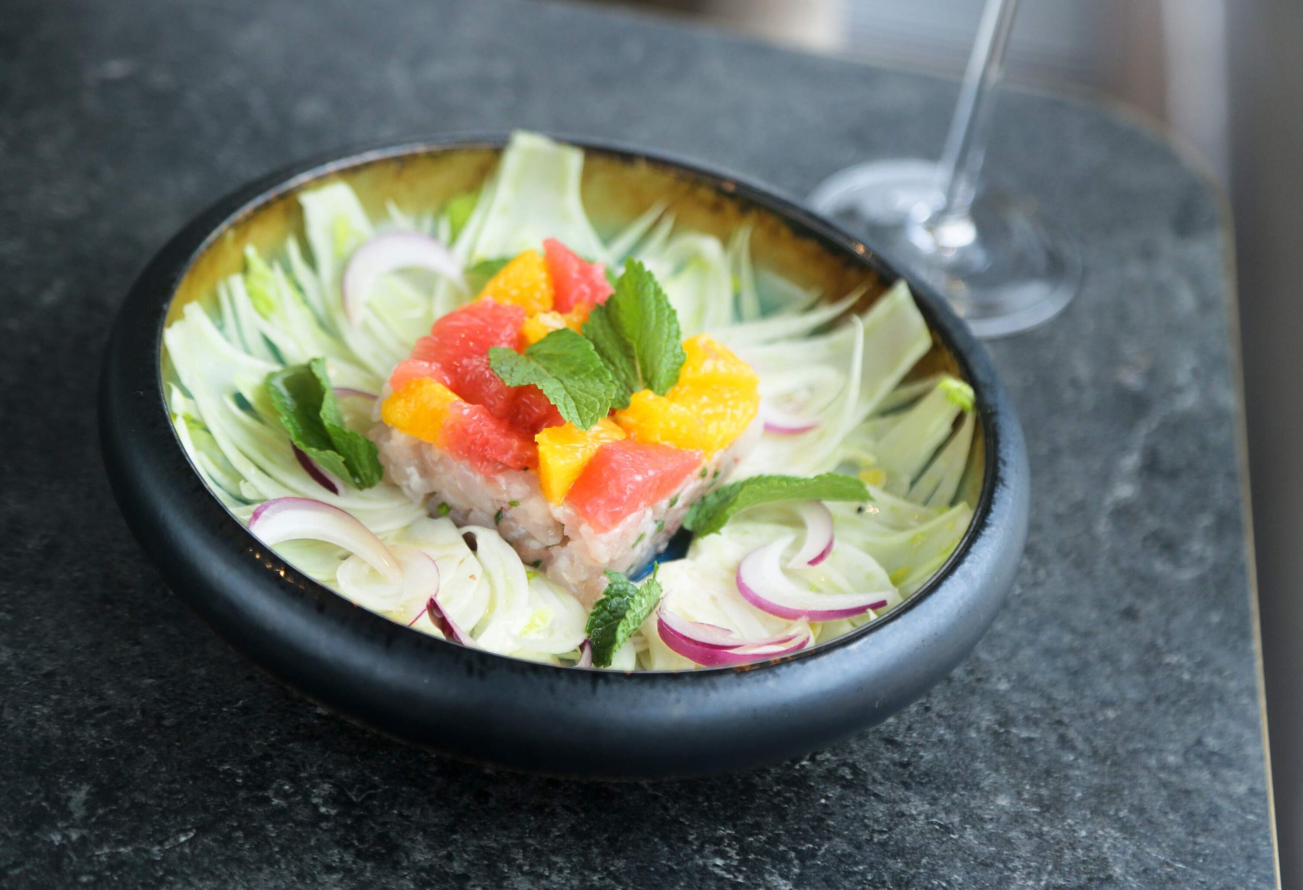 A bowl of raw fish dish garnished with onion, mint leaves, and orange slices.