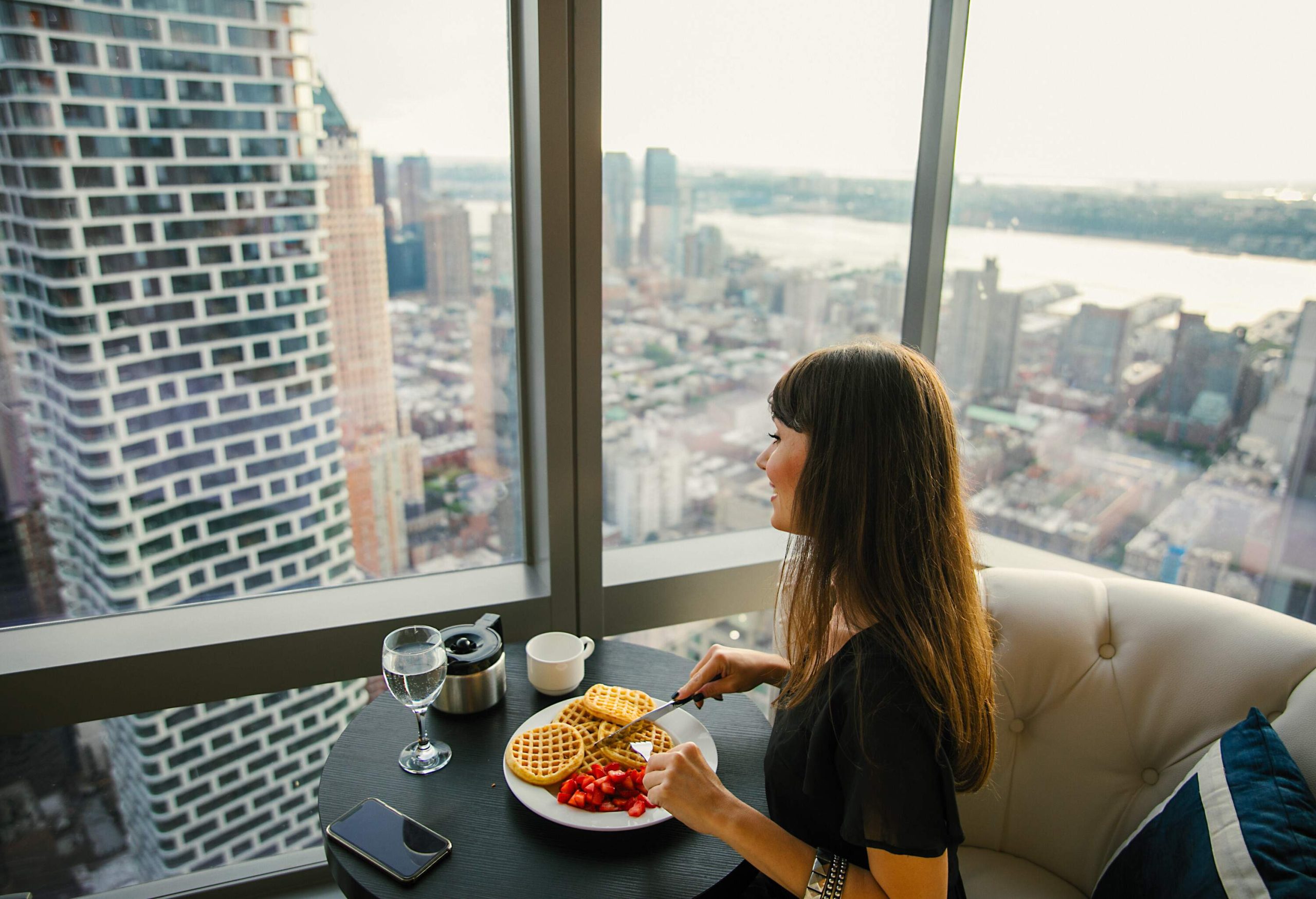 A woman eating waffles and strawberries at a table while she looks out over the city through the glass windows.