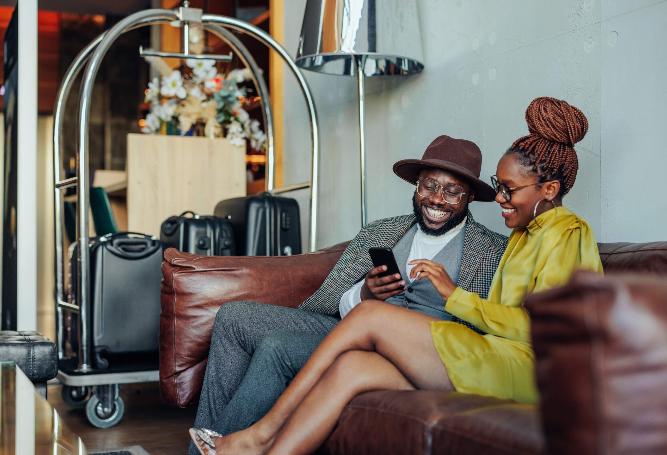 Couple sitting on a sofa in hotel lobby with their luggage on the cart, using a smartphone.