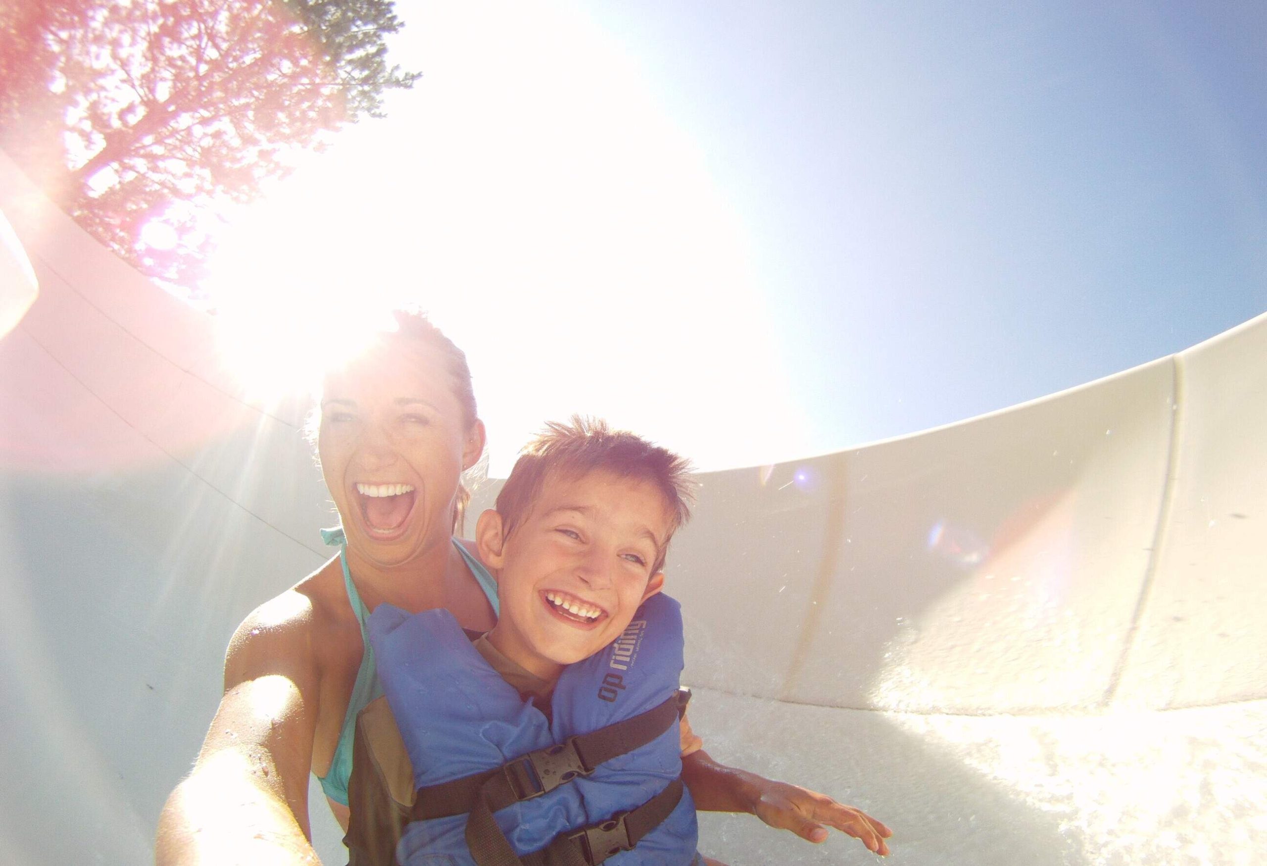 A woman with a kid wearing a life jacket smile as they slide down a water slide.