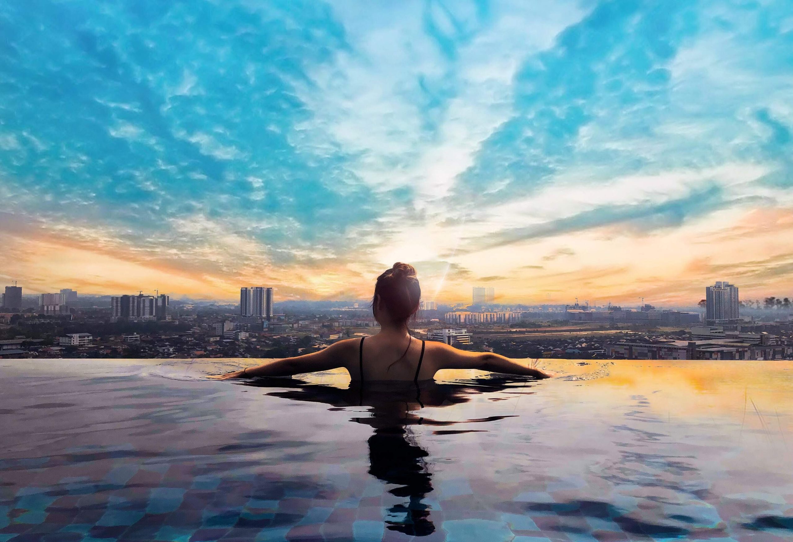 A rear view of a woman at the edge of an infinity pool with a cityscape view against the dramatic sky.
