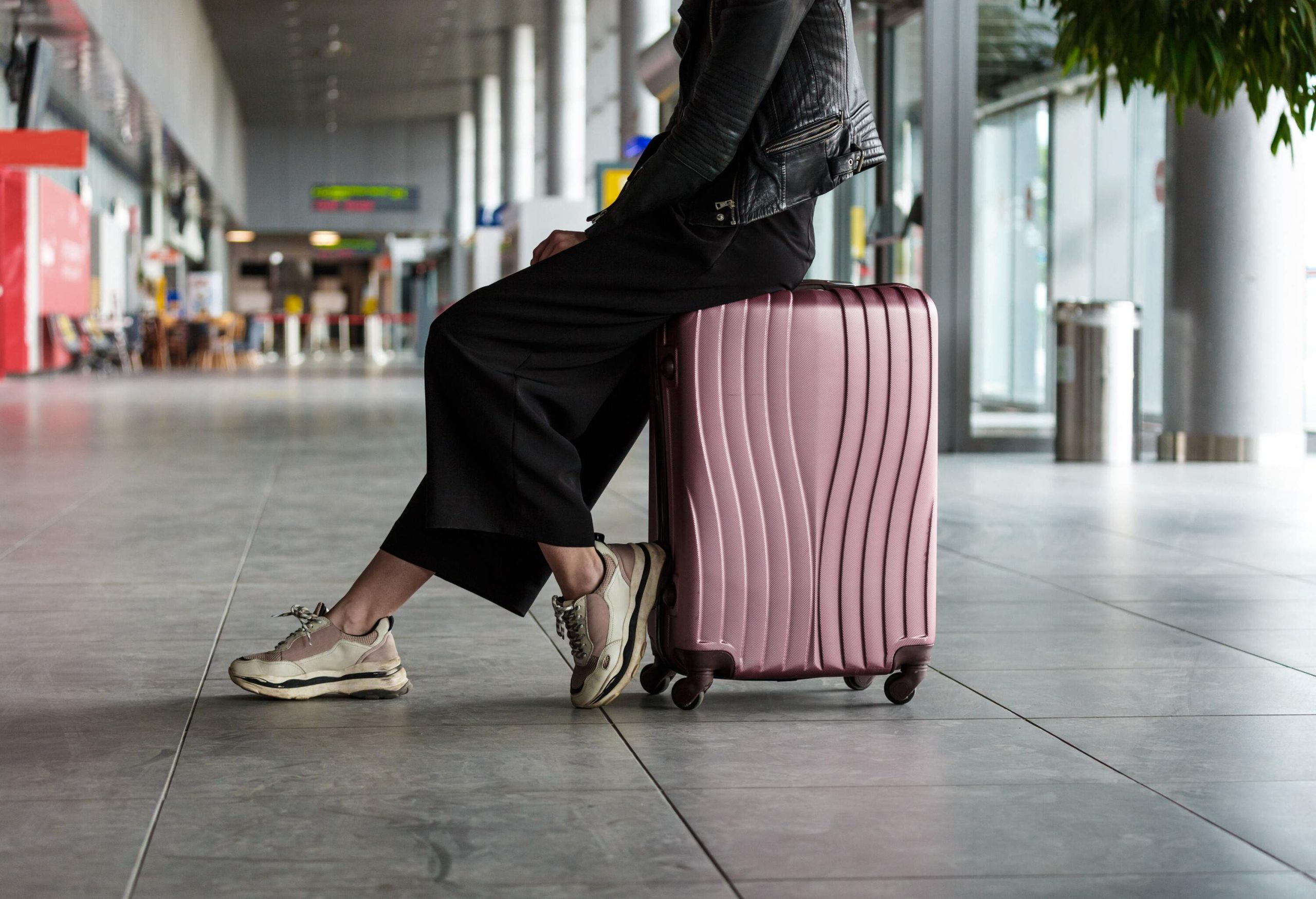 A woman in a black skirt sits on a silver-coloured luggage in an airport lobby.