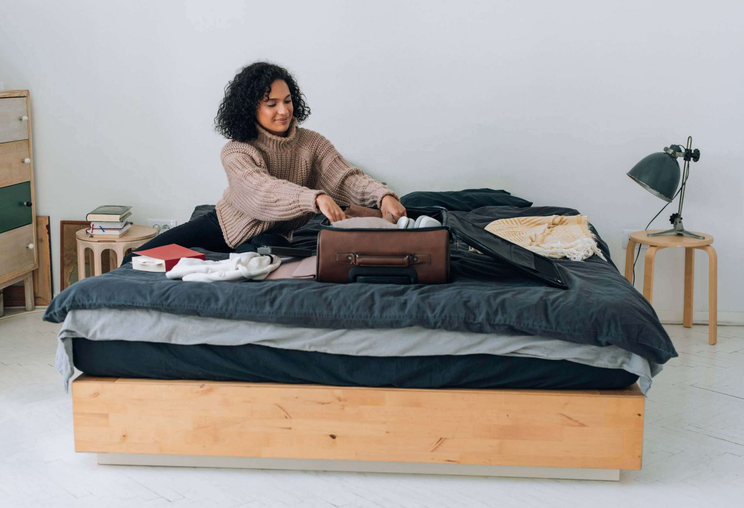 A curly-haired lady fixing her clothes into a brown suitcase on the bed.