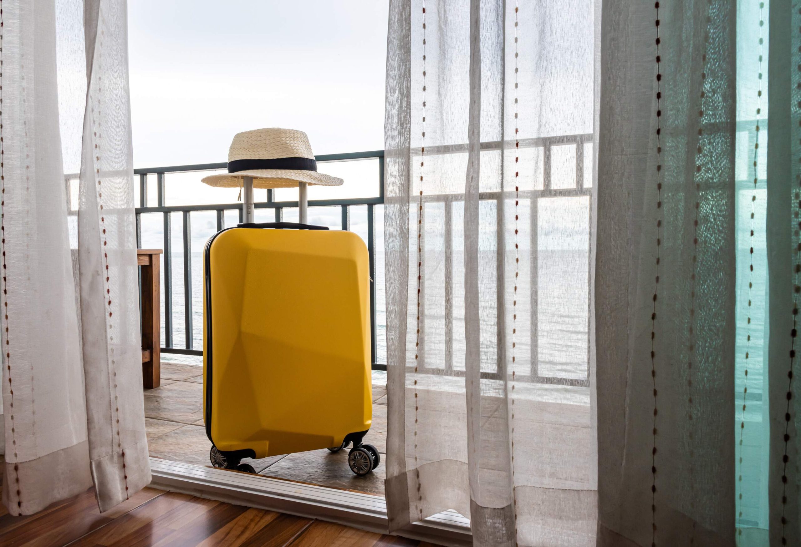A bright yellow suitcase with a hat on its handle standing on a patio behind the glass doors.