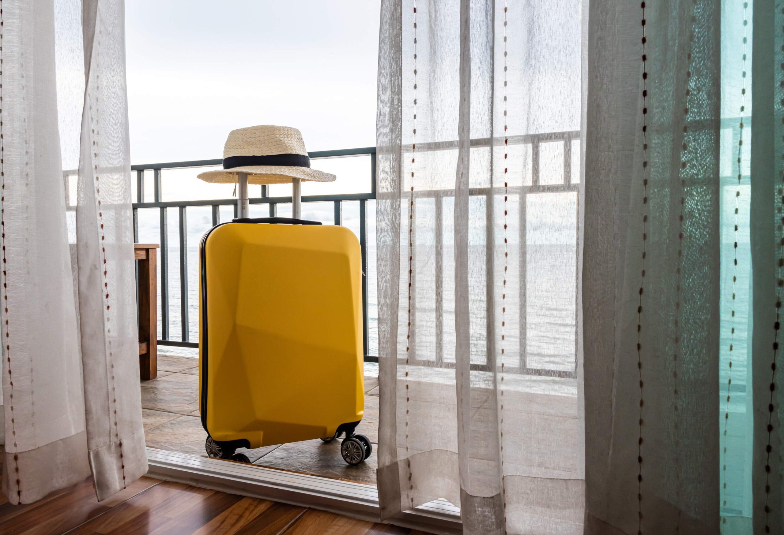 A bright yellow suitcase with a hat on its handle standing on a patio behind the glass doors.