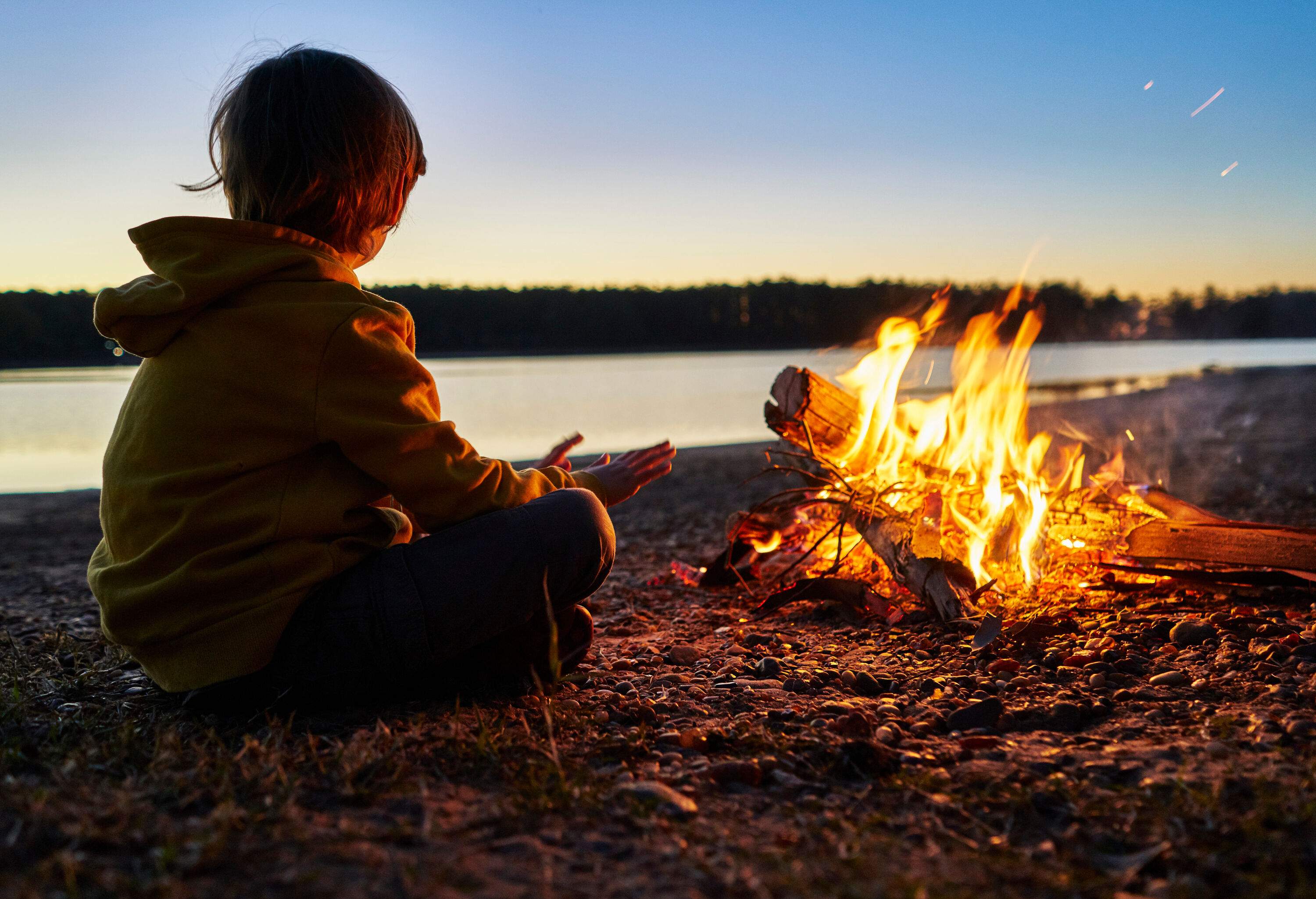 A child in a hooded jacket sits cross-legged in front of a bonfire with open hands.