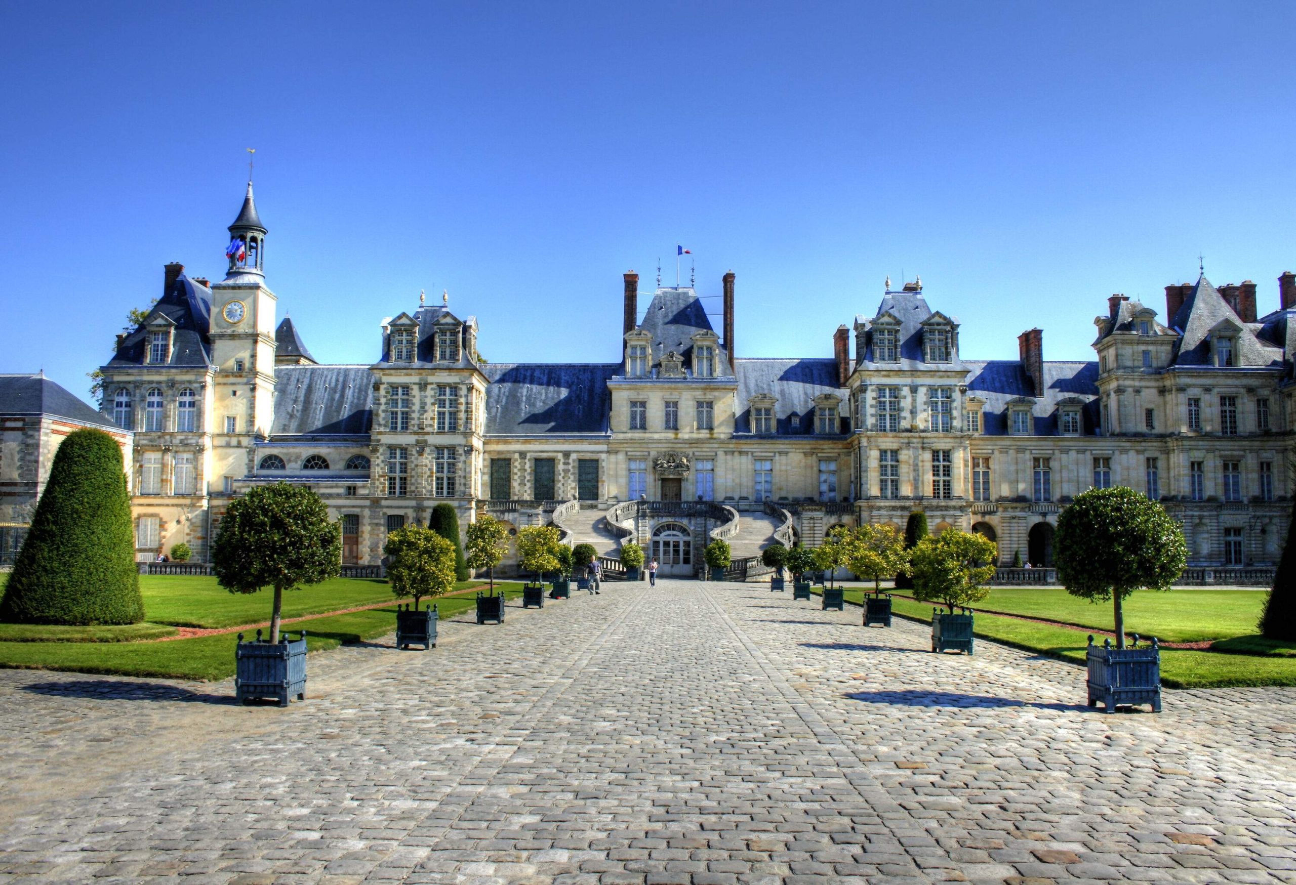 A cobblestone path lined with potted plants and green lawn leads to the Palace of Fontainebleau, a medieval manor house with an outdoor double staircase, multiple windows, and blue roofing.