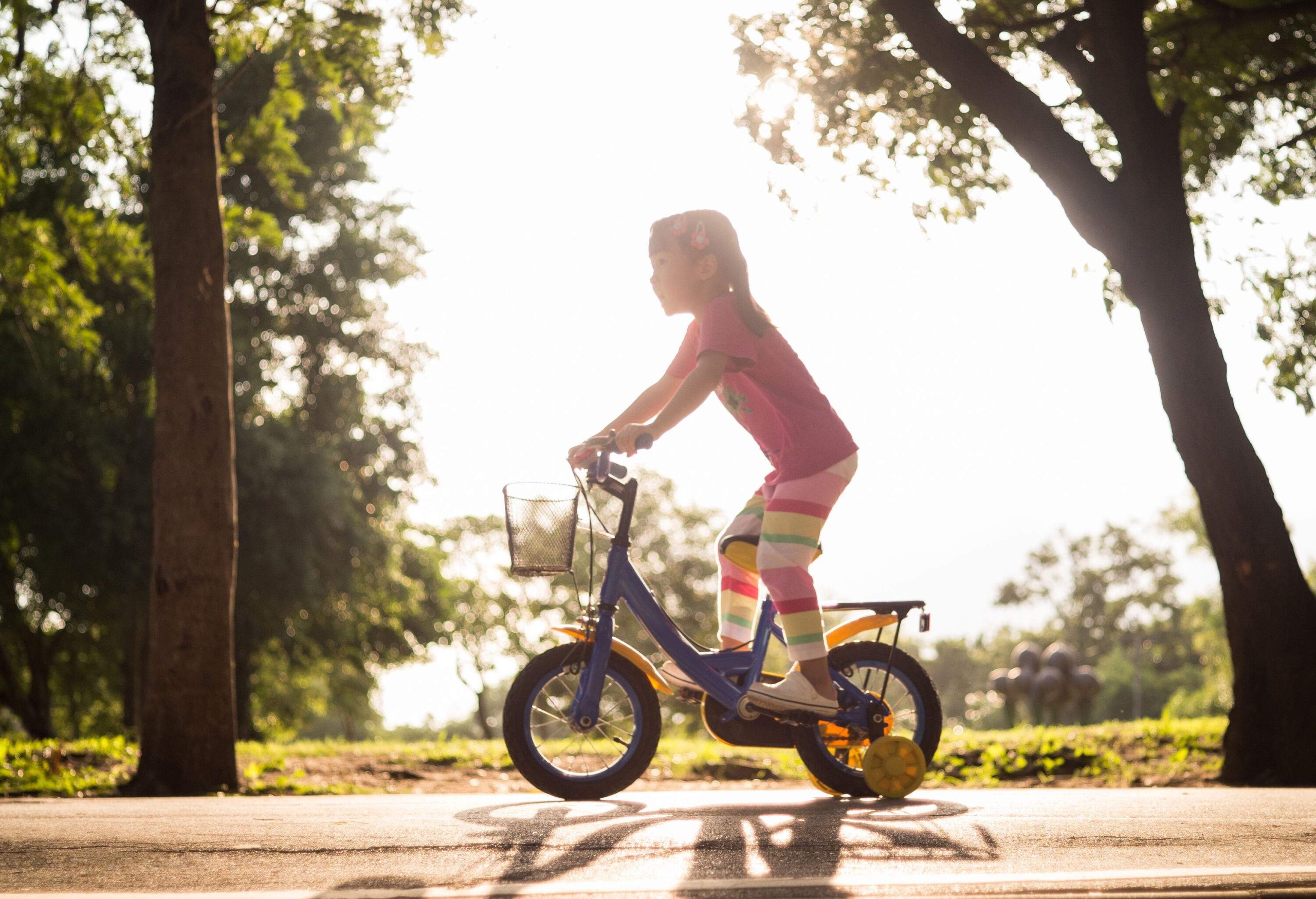 A girl rides her tiny bike on a sunlit asphalt road lined with tall trees.