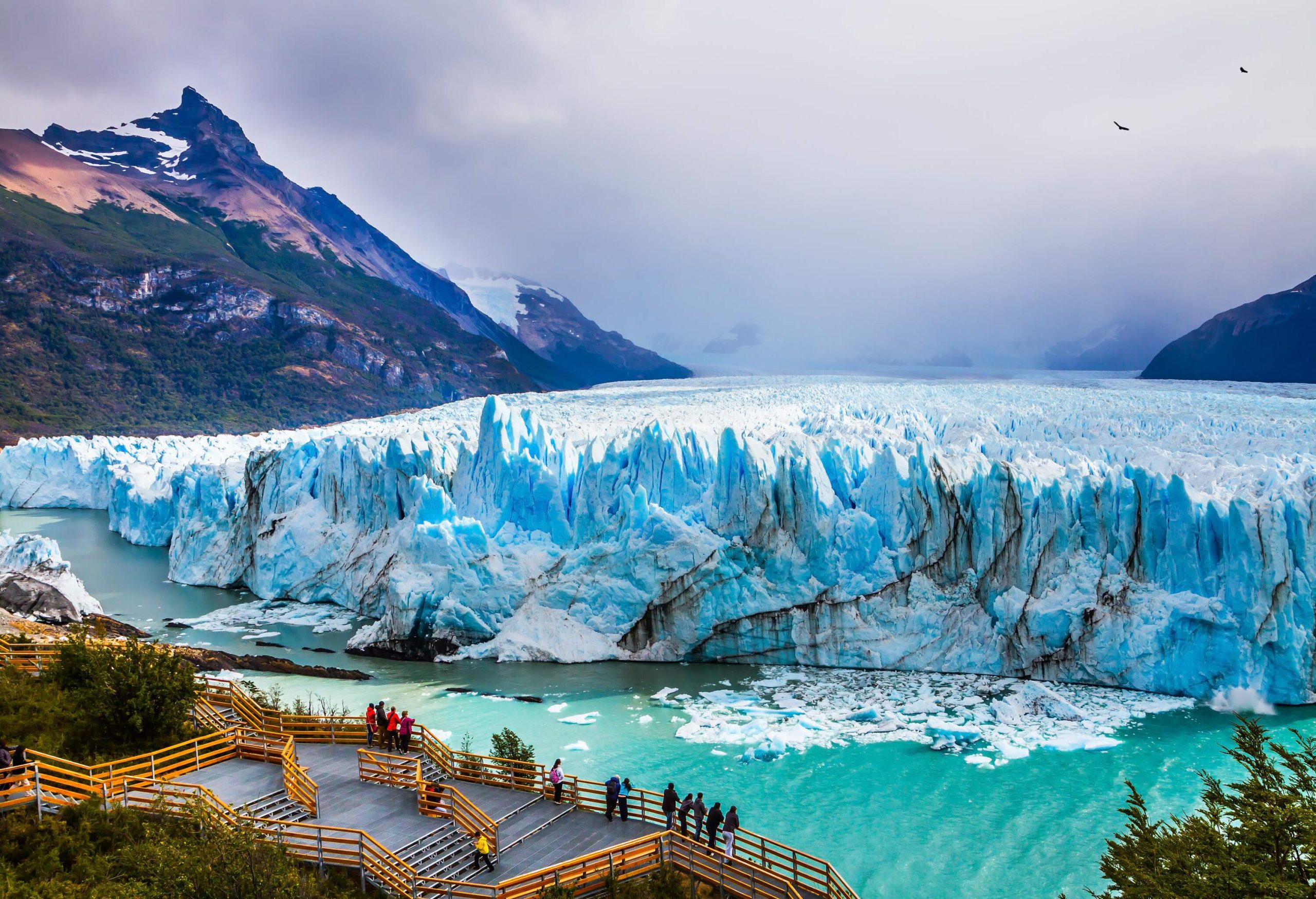 People on a boardwalk watching a glacier gradually melting into a lake.