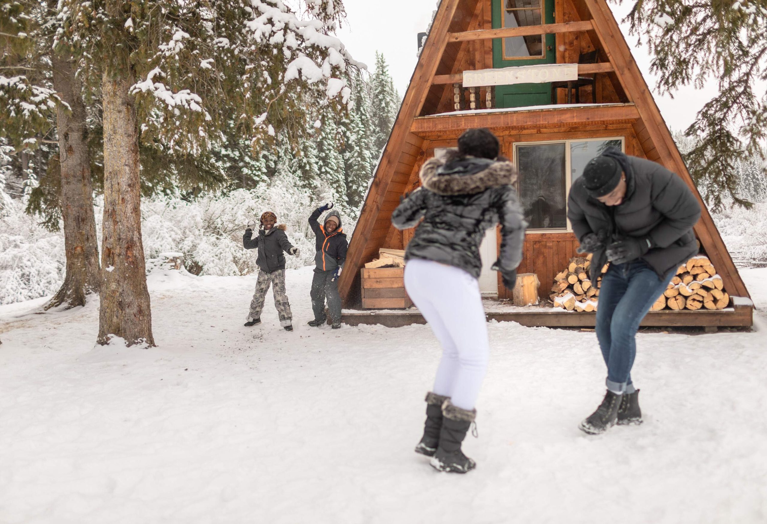 A trendy couple is caught off guard by two playful children, unleashing a flurry of snowballs outside their charming A-frame holiday cabin.