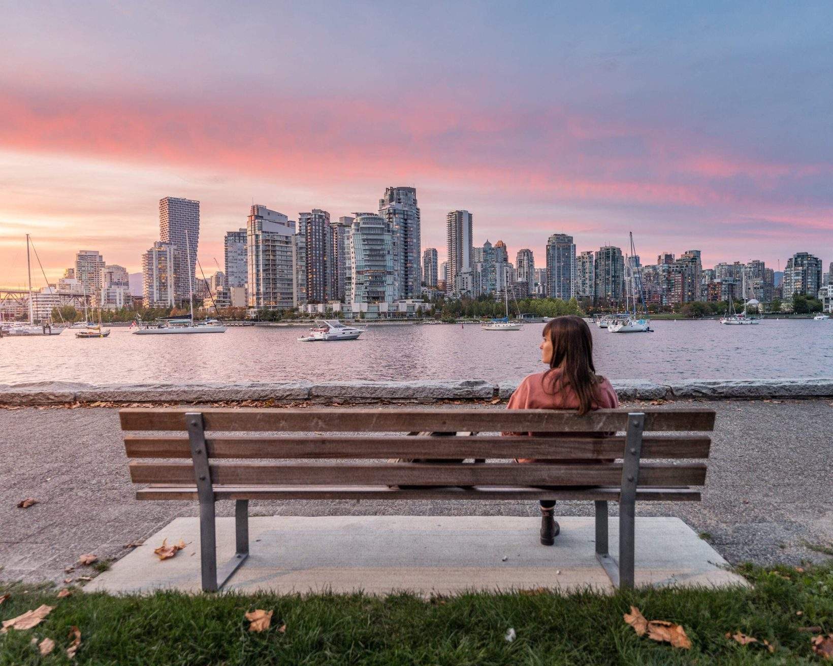 A woman sits on a bench in front of a harbour full of cruising boats with distant views of the city skyline.