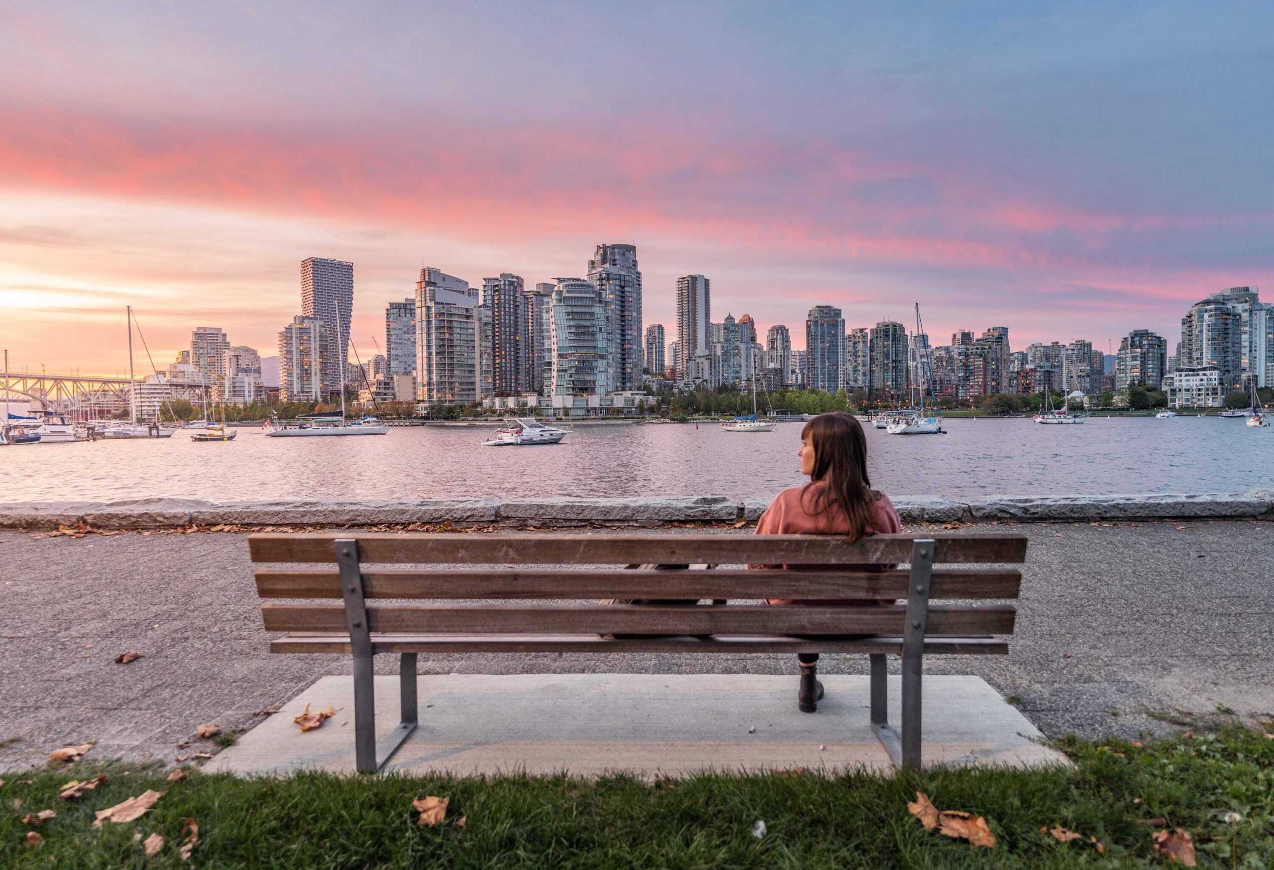 A woman sits on a bench in front of a harbour full of cruising boats with distant views of the city skyline.