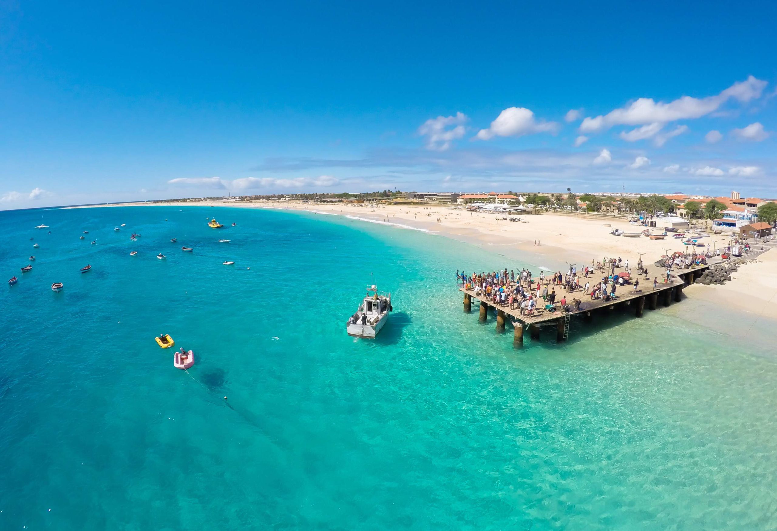 A crowded pier on a turquoise beach with floating boats.