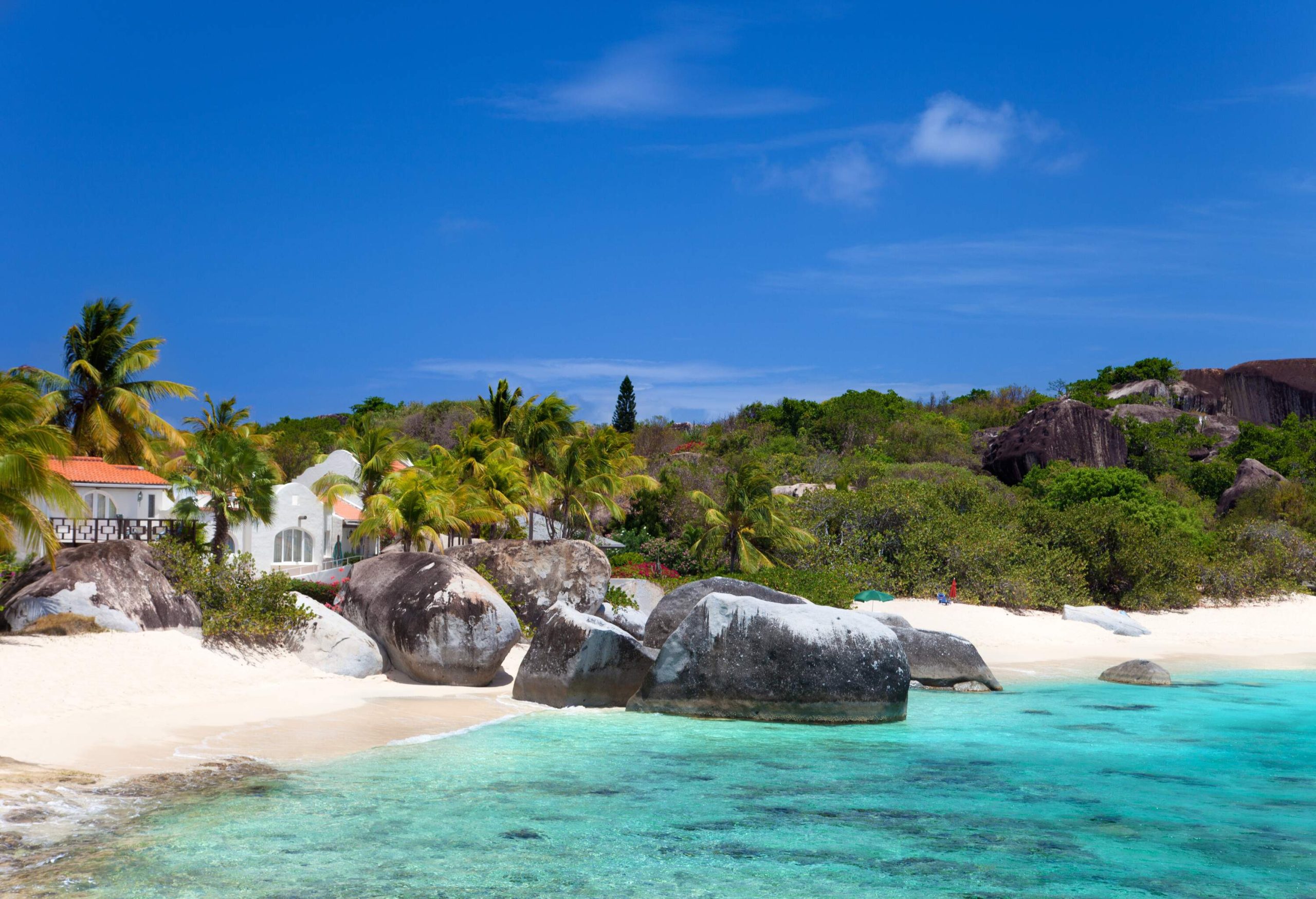 Huge boulders scattered along the white sand shore, with beach houses nestled amidst tall green trees, all surrounded by the captivating turquoise sea.