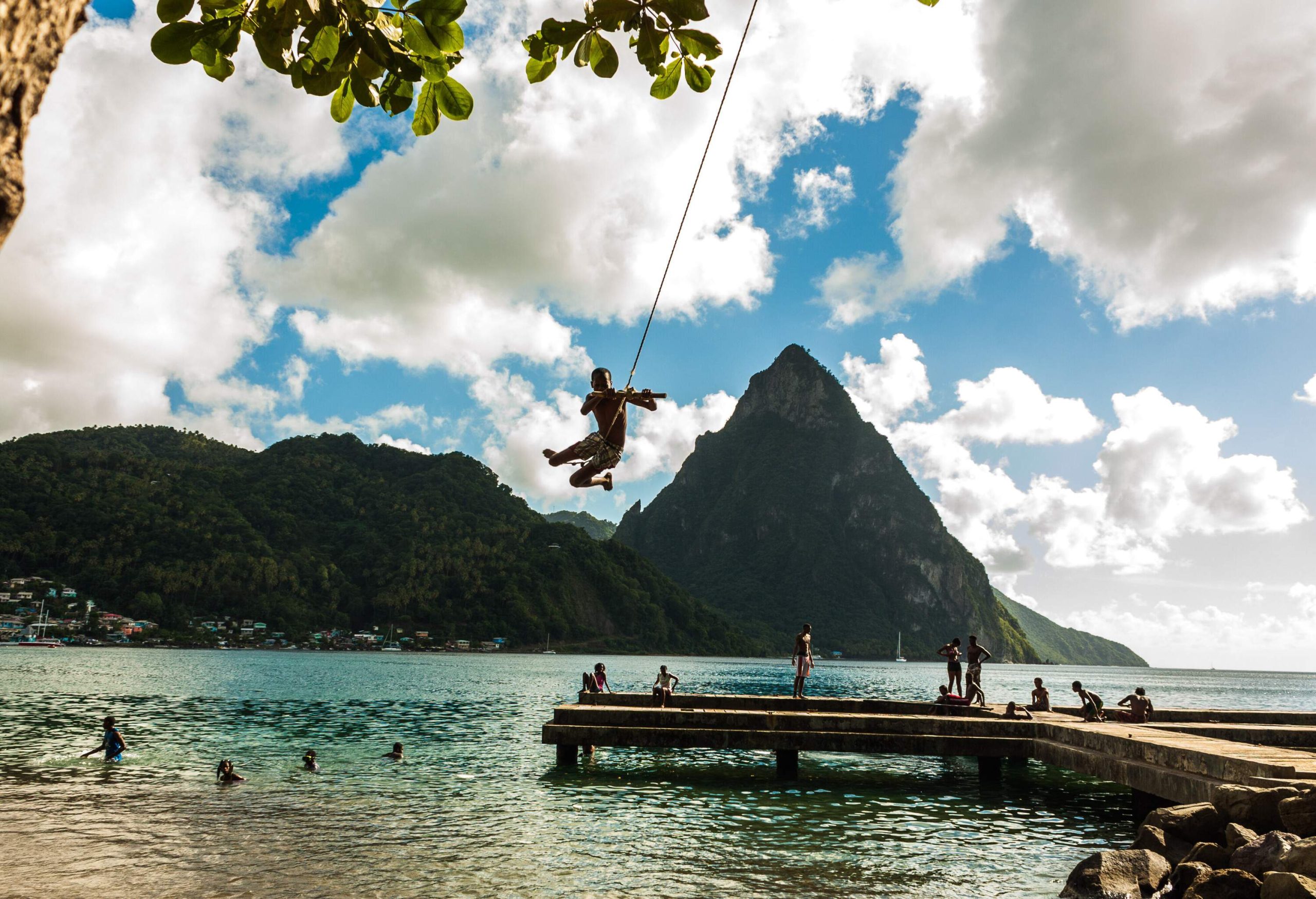 A person jumps from a rope swing into the water as a group of people watch from a concrete pier.