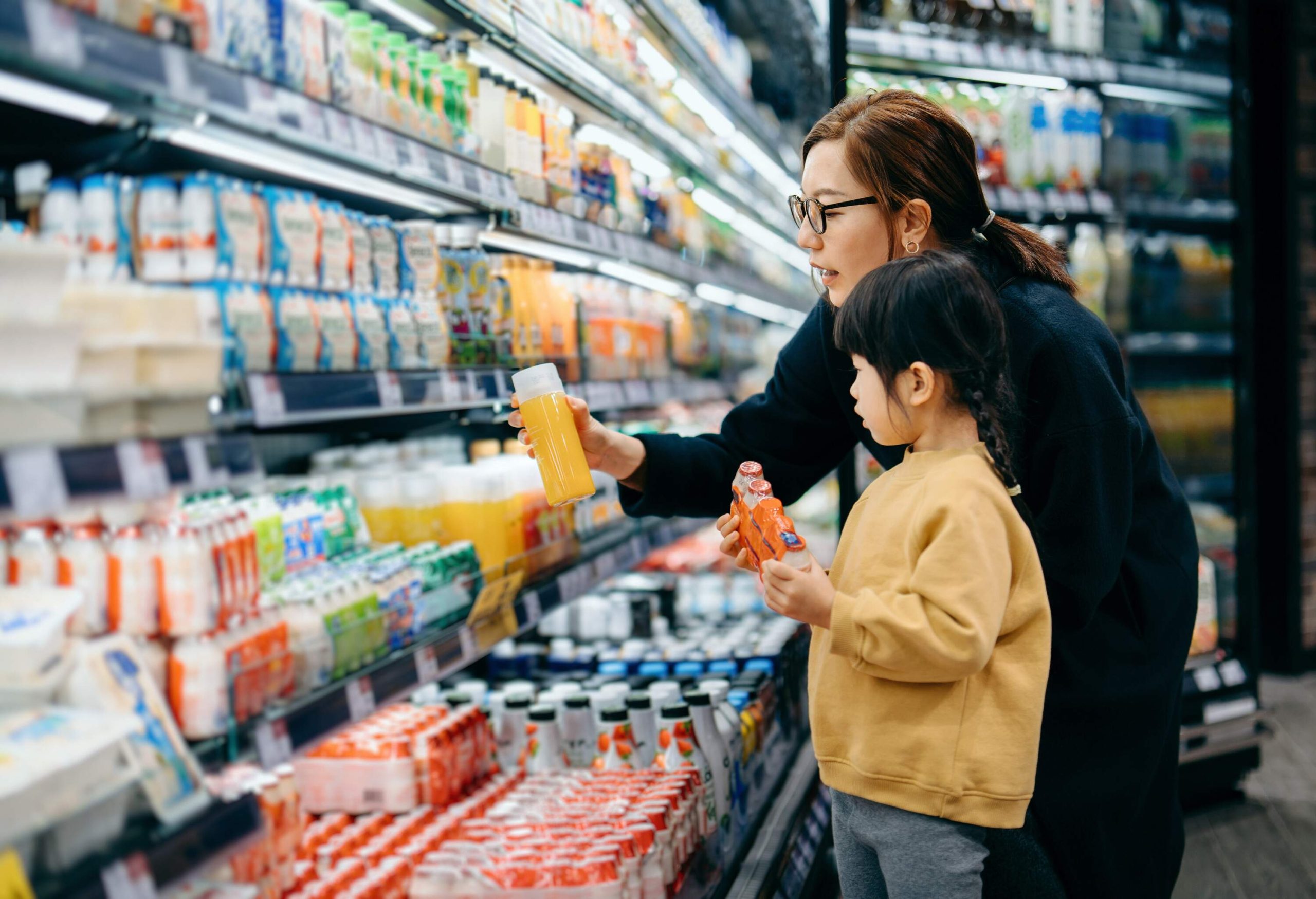 Young Asian mother and her little daughter grocery shopping in supermarket. they are choosing fresh fruit juice together along the beverage aisle. Routine grocery shopping. Healthy eating lifestyle.