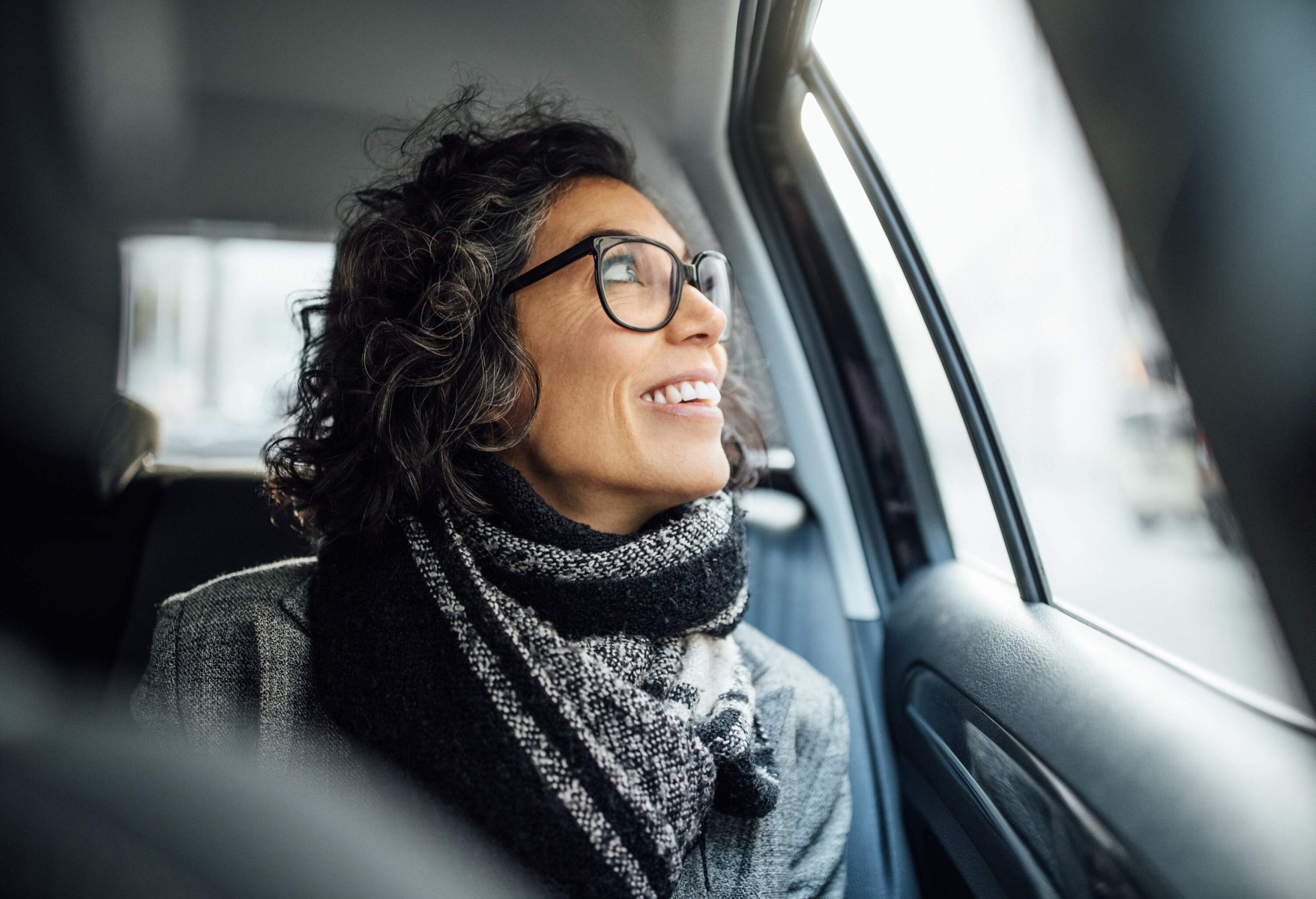 Mature woman sitting in backseat of a car looking out the window and smiling. Mid adult female traveling by a car.