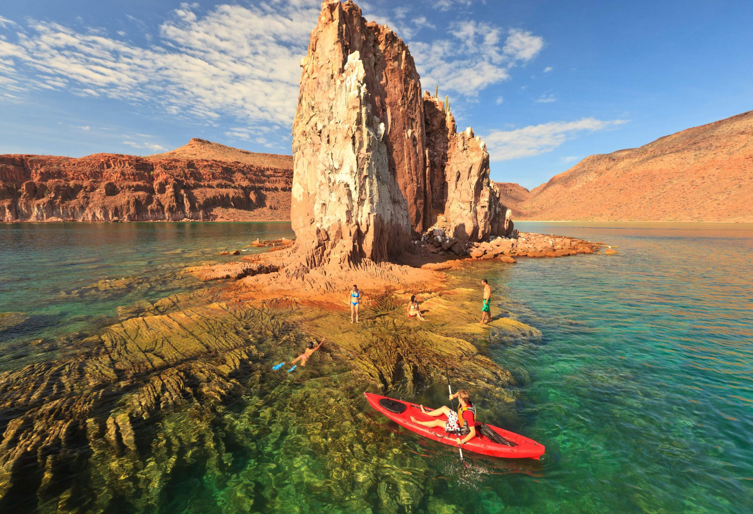 A man in a red canoe watching the people on the shore of a rock island.