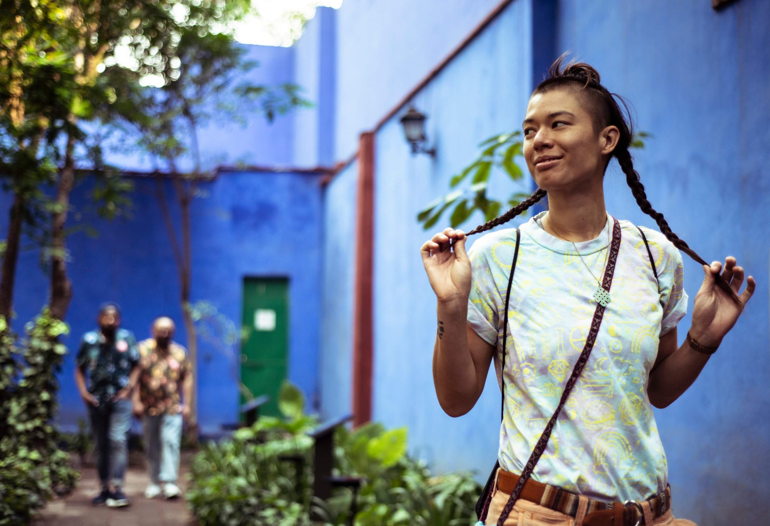 A young woman standing in the yard of a blue-walled building holding her braided hair, with two men walking on a path behind her.