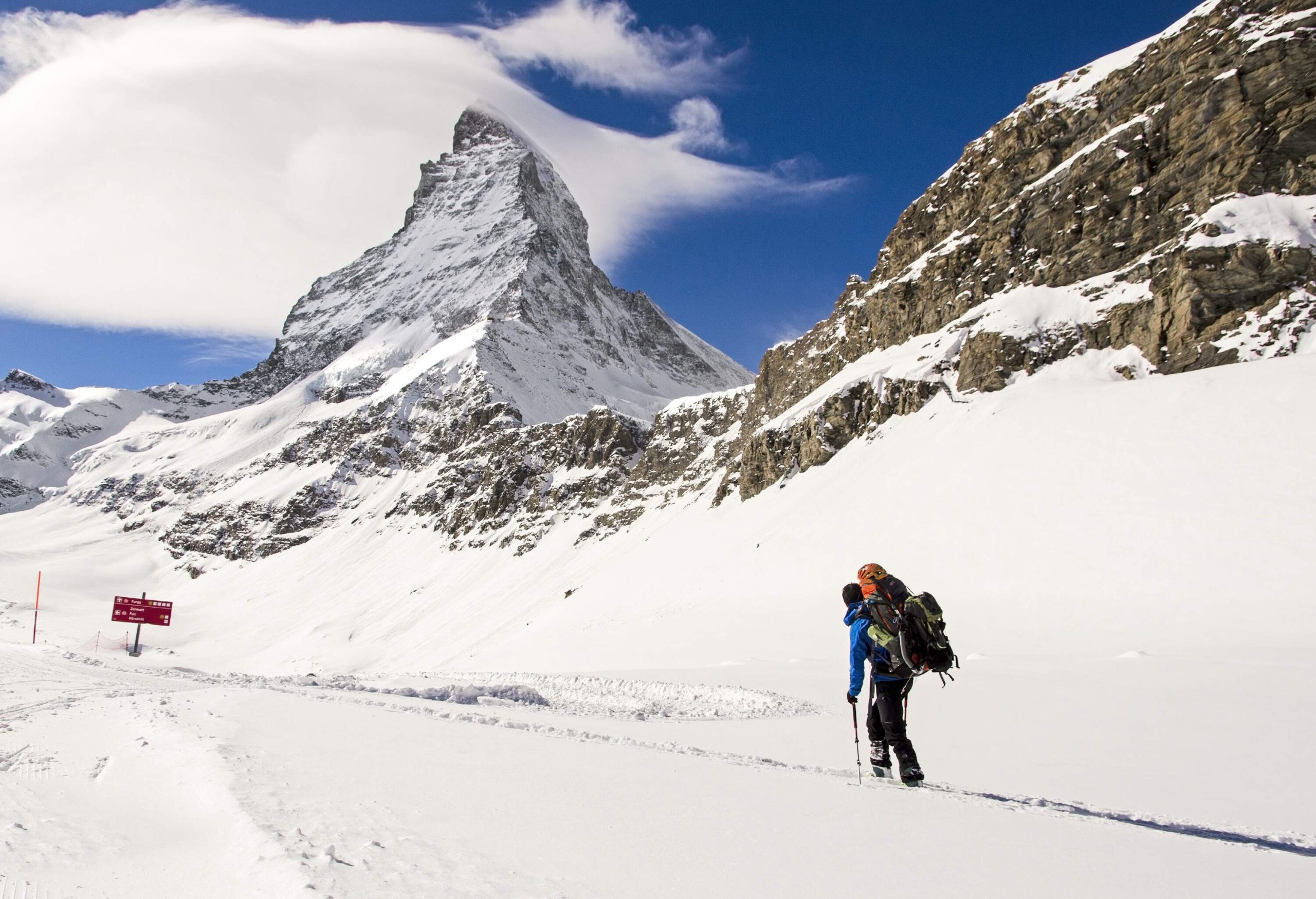 A lone traveller in full winter gear carry a bag while walking across a snow trail along the snow-capped mountain range.