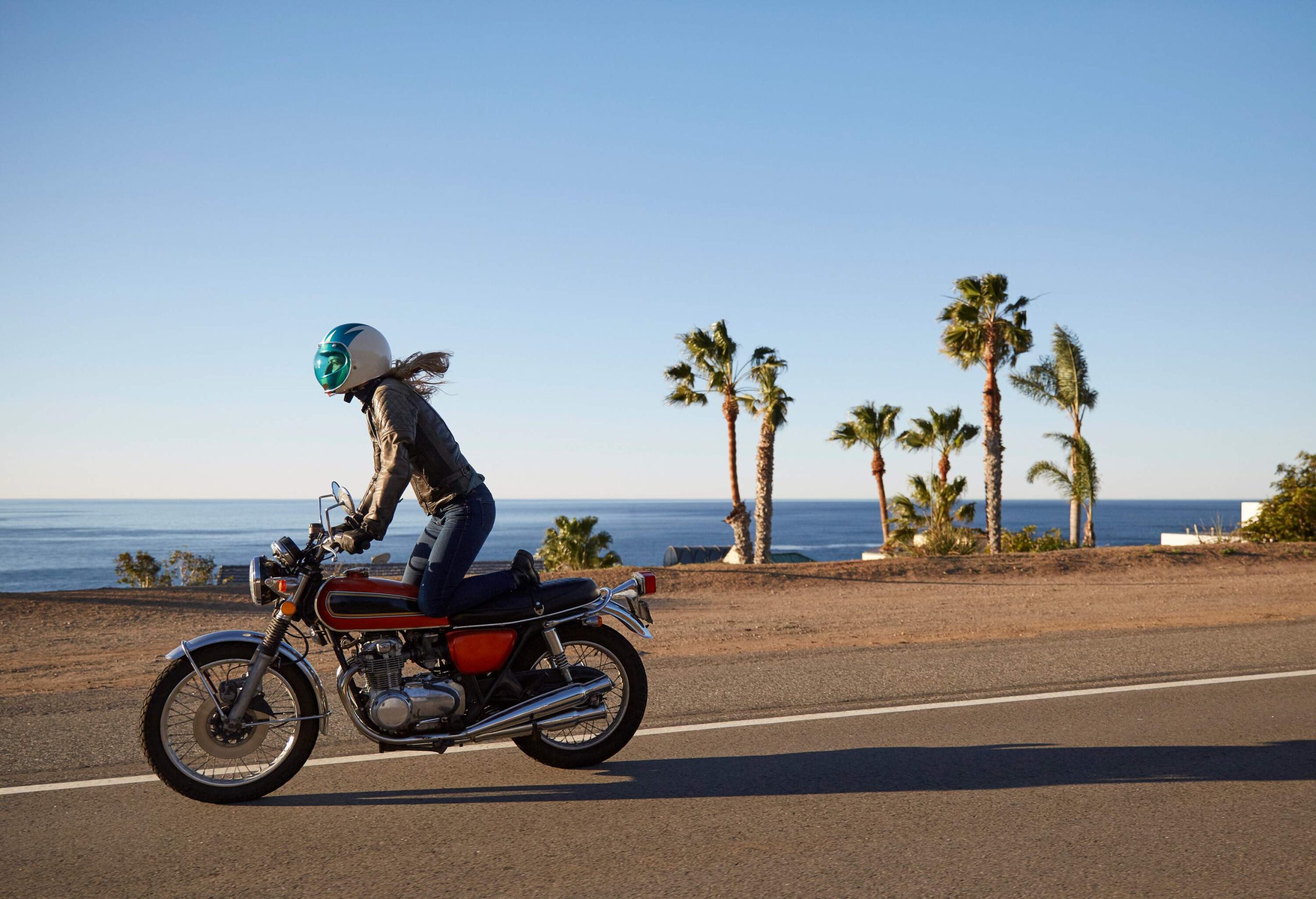 A woman in motor riding gear enjoys riding a motorbike along a paved road that runs alongside the captivating sea.