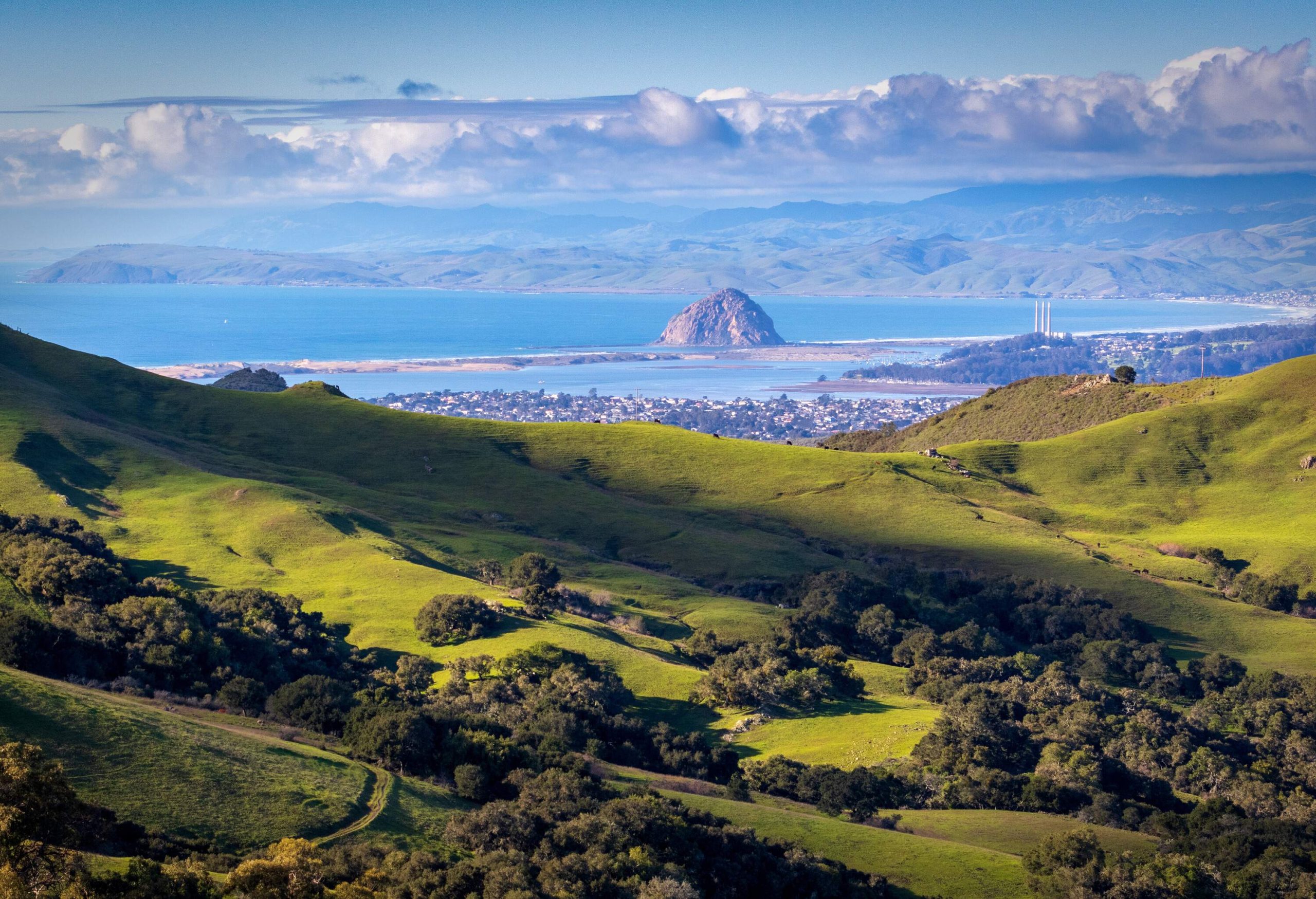 A massive rock in the centre of a bay near a coastal village as seen from sloping hills covered in greenery.