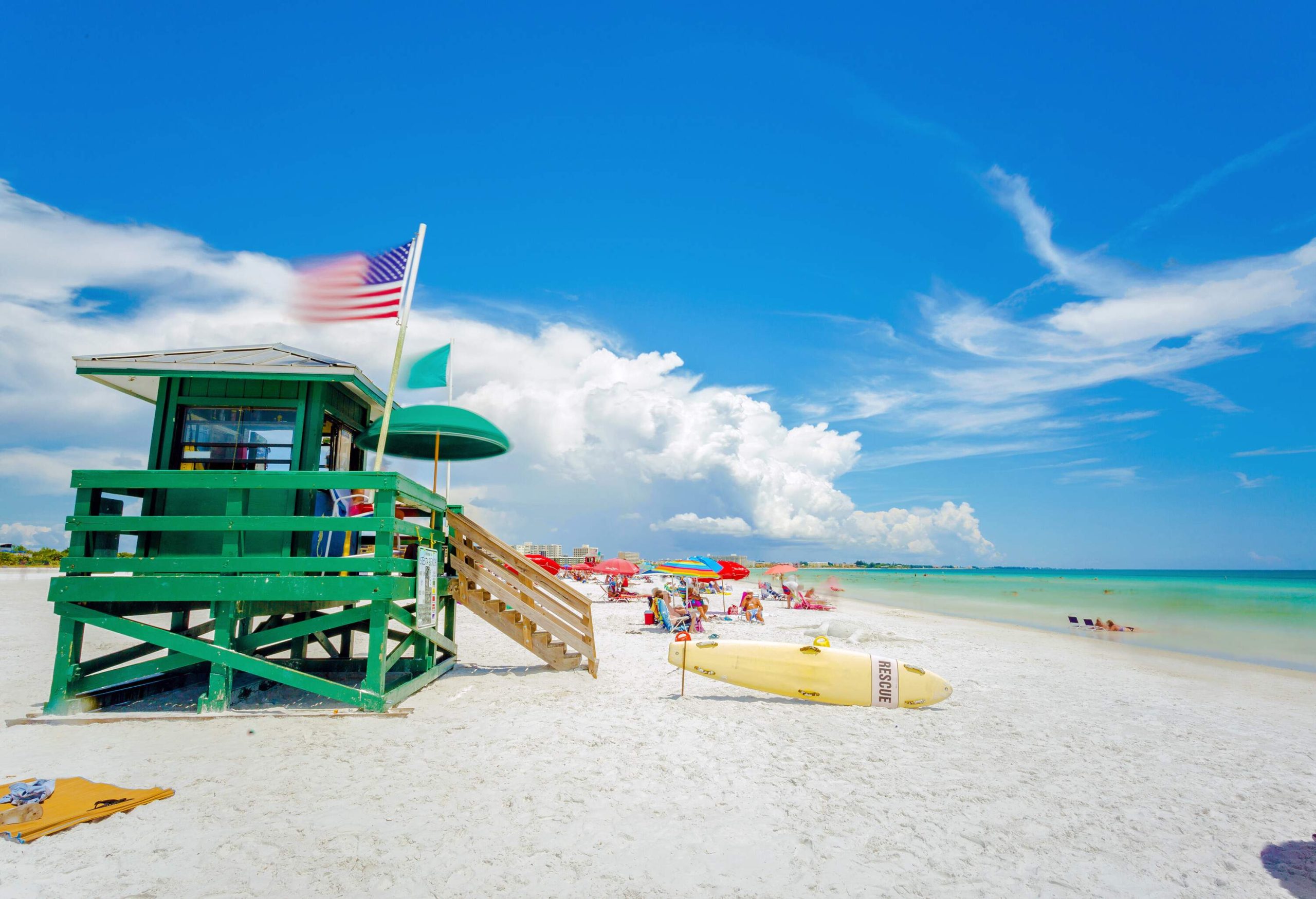 A lifeguard tower next to a yellow surfboard on a white beach with people lounging on sunbeds and umbrellas.