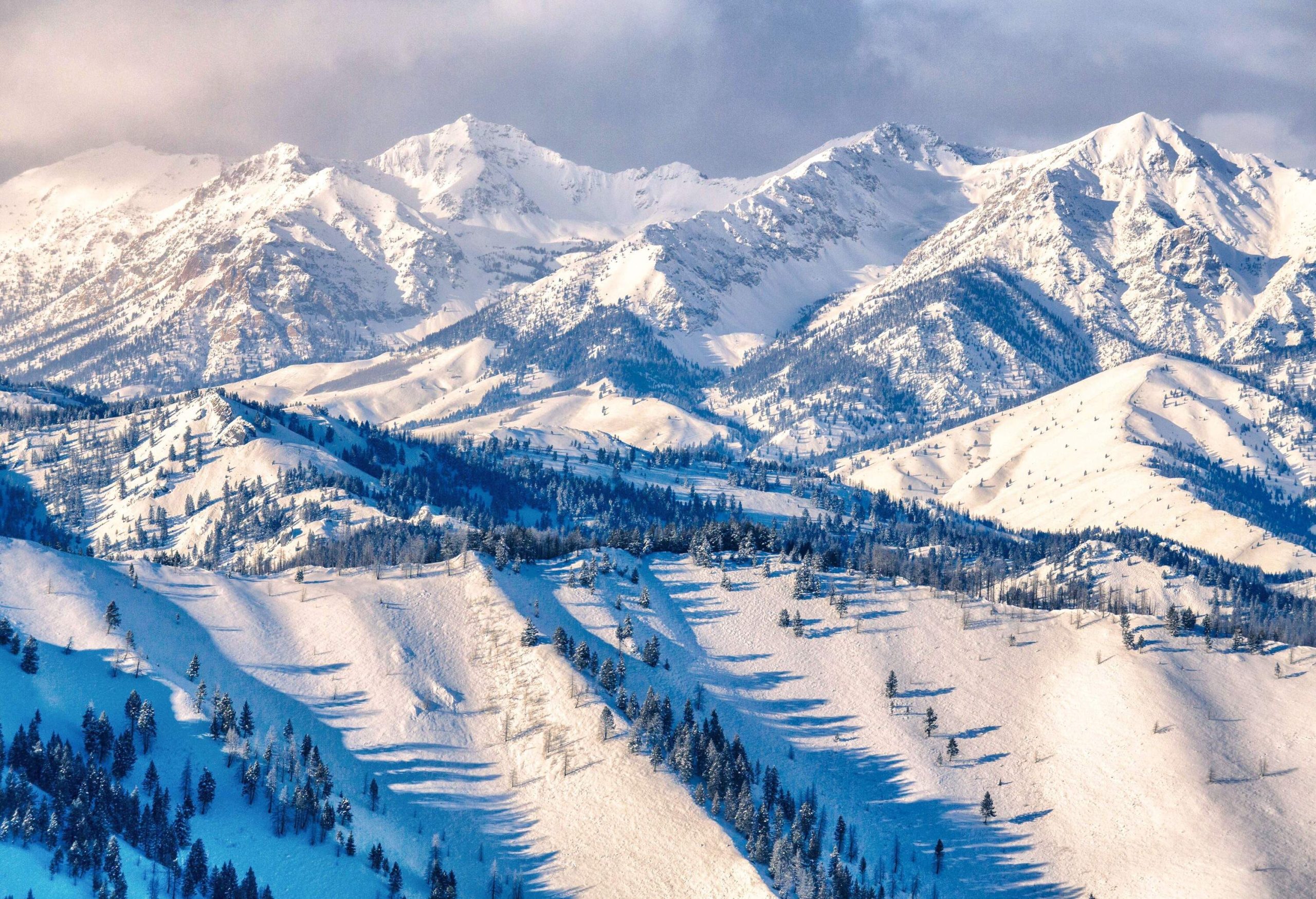 The landscape of sloppy ski terrain lined with frosted tall trees beneath the steep rocky mountains covered in deep snow.