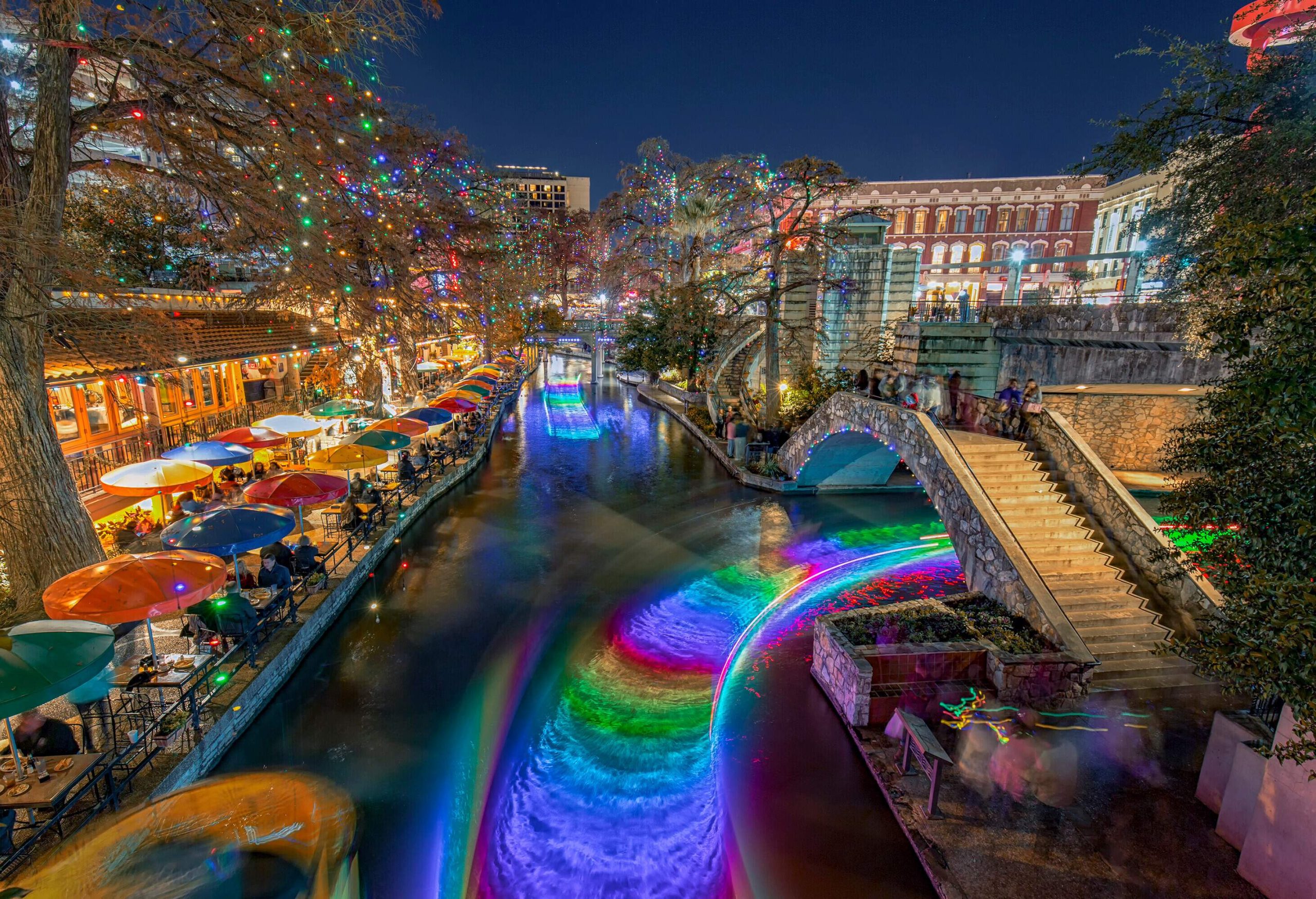 A popular esplanade with outdoor chairs and tables runs alongside a canal with rainbow water splash.