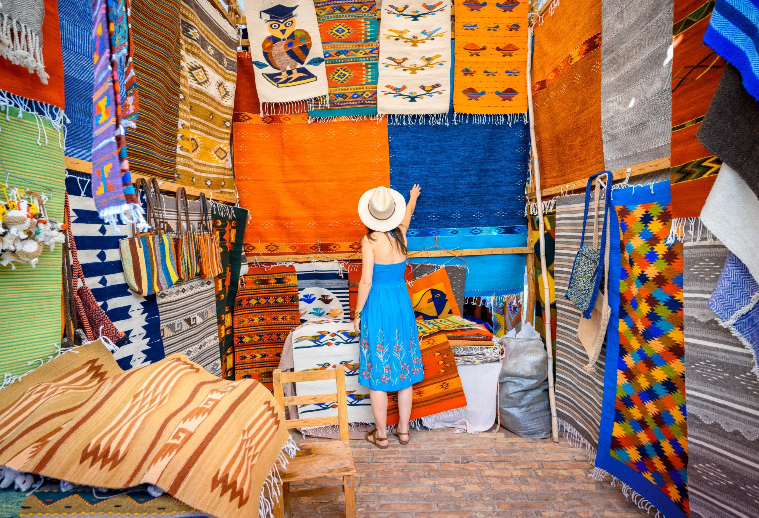 A curious child, dressed in a blue dress and sunhat, gazes in wonder at the array of carpets in a store, eagerly reaching out to touch the one directly in front of them.