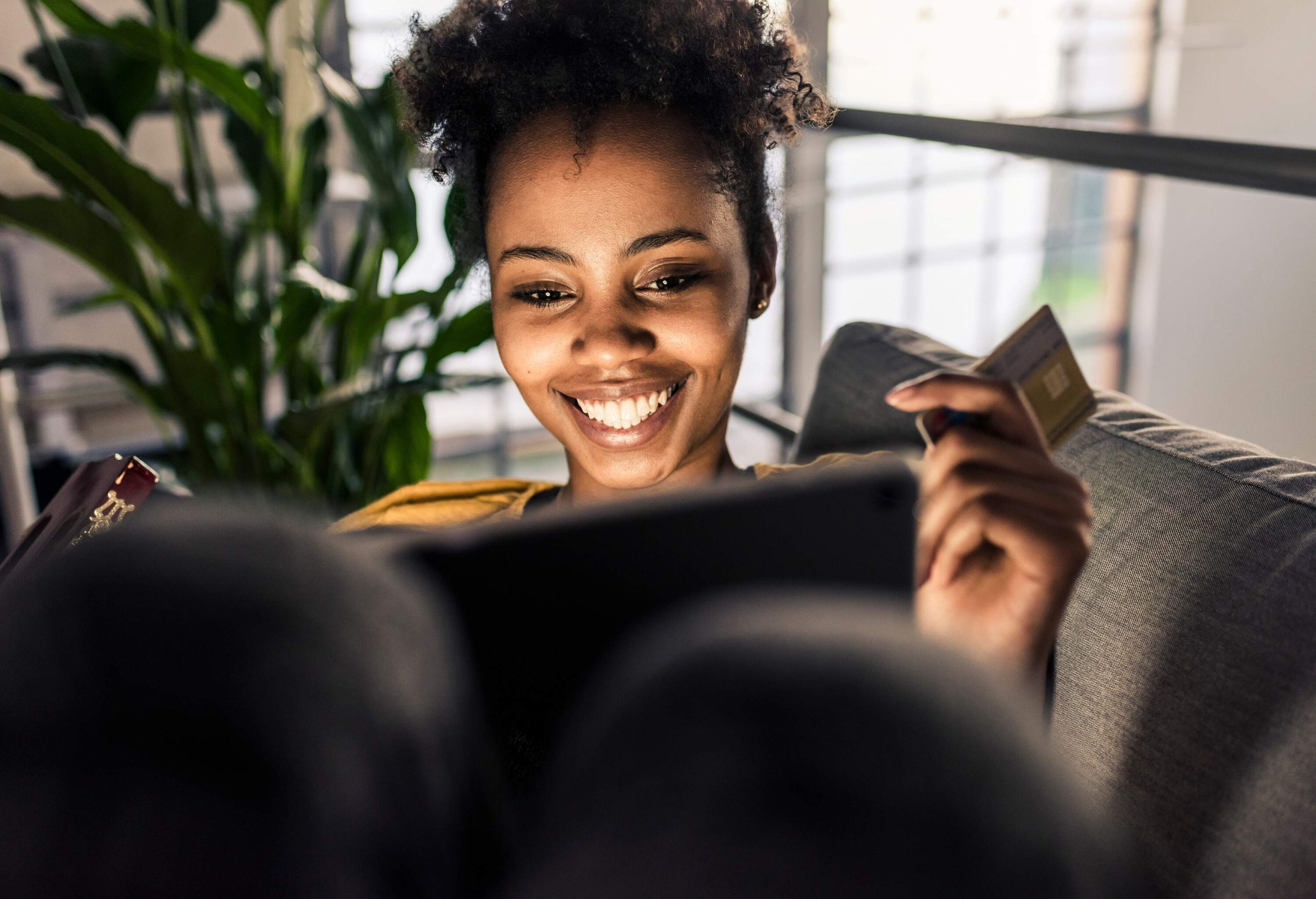 A curly-haired woman sits on a sofa and smiles at her laptop while holding a credit card.