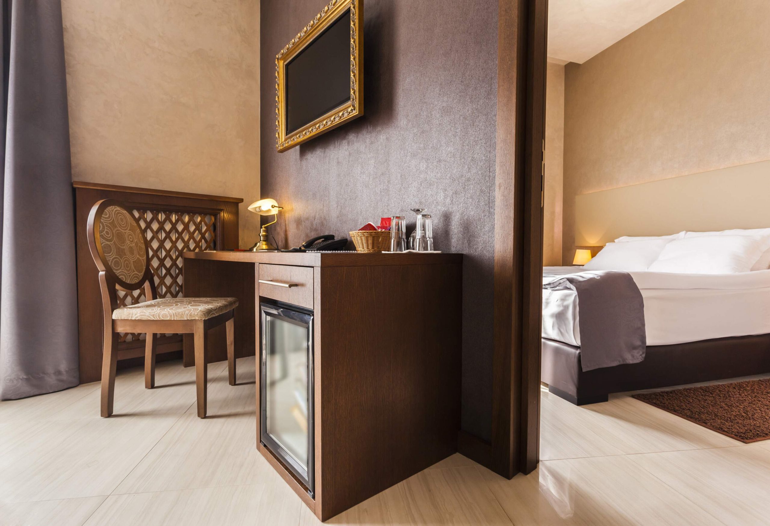 A hotel room with a dark wood work desk separated by a partition wall from a sizeable bed covered in clean linens.