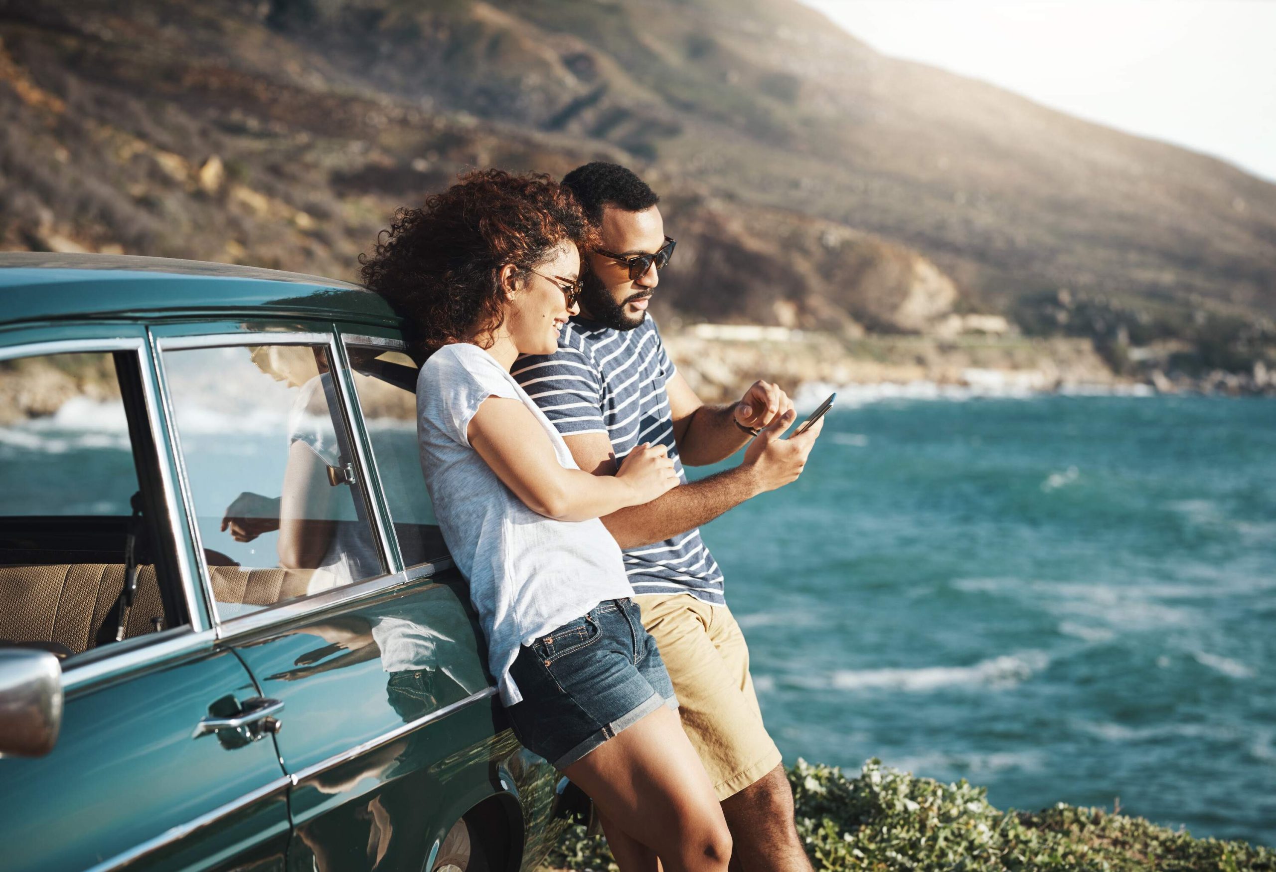 A couple in casual clothes looks at a phone while leaning on a black car beside a beach.