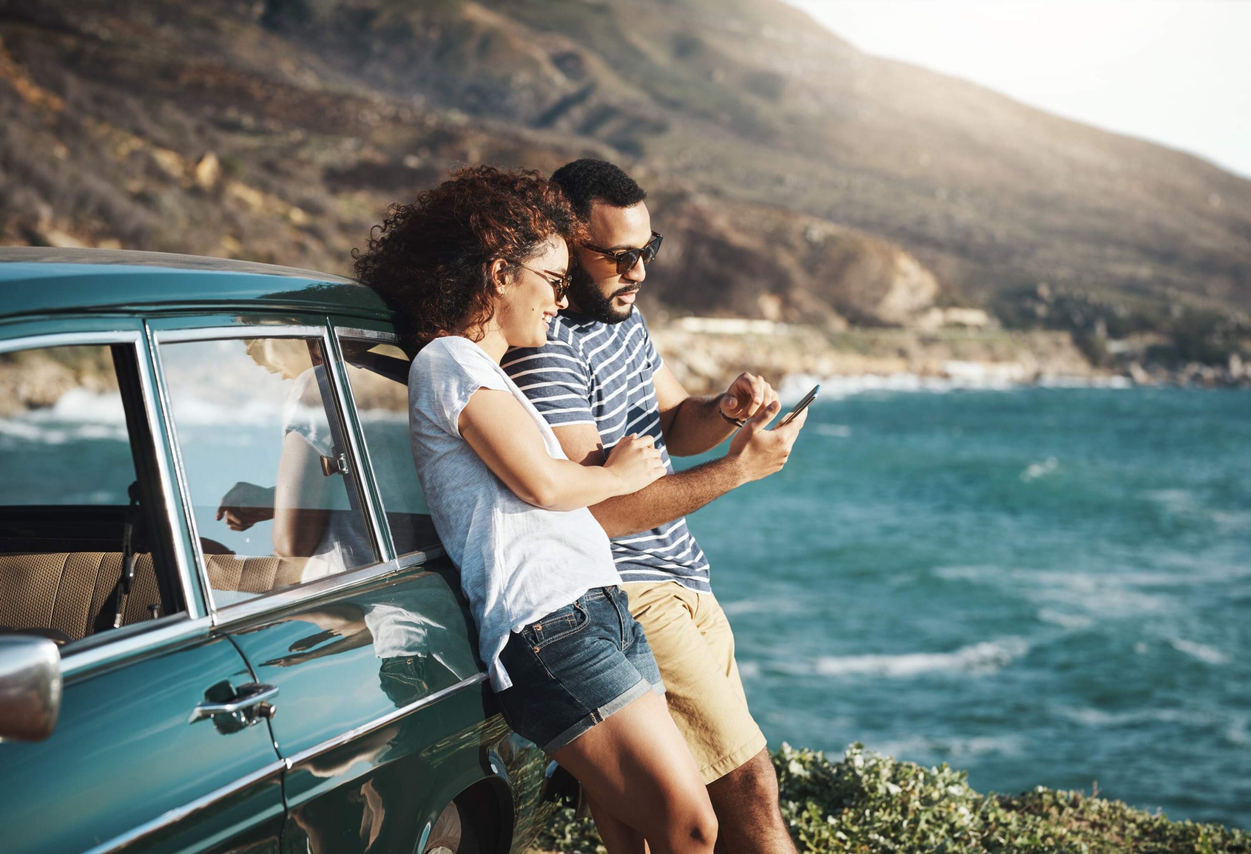 A couple in casual clothes looks at a phone while leaning on a black car beside a beach.
