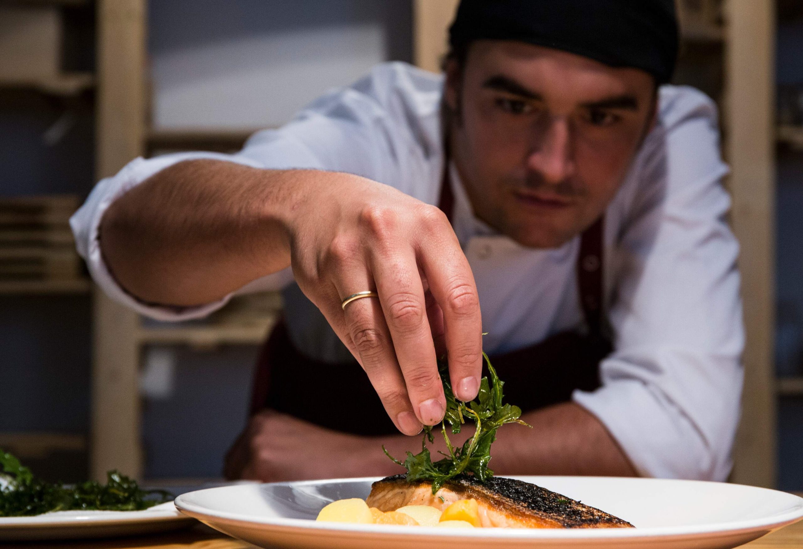 A male chef garnishes the slice of salmon with sauteed green leaves.