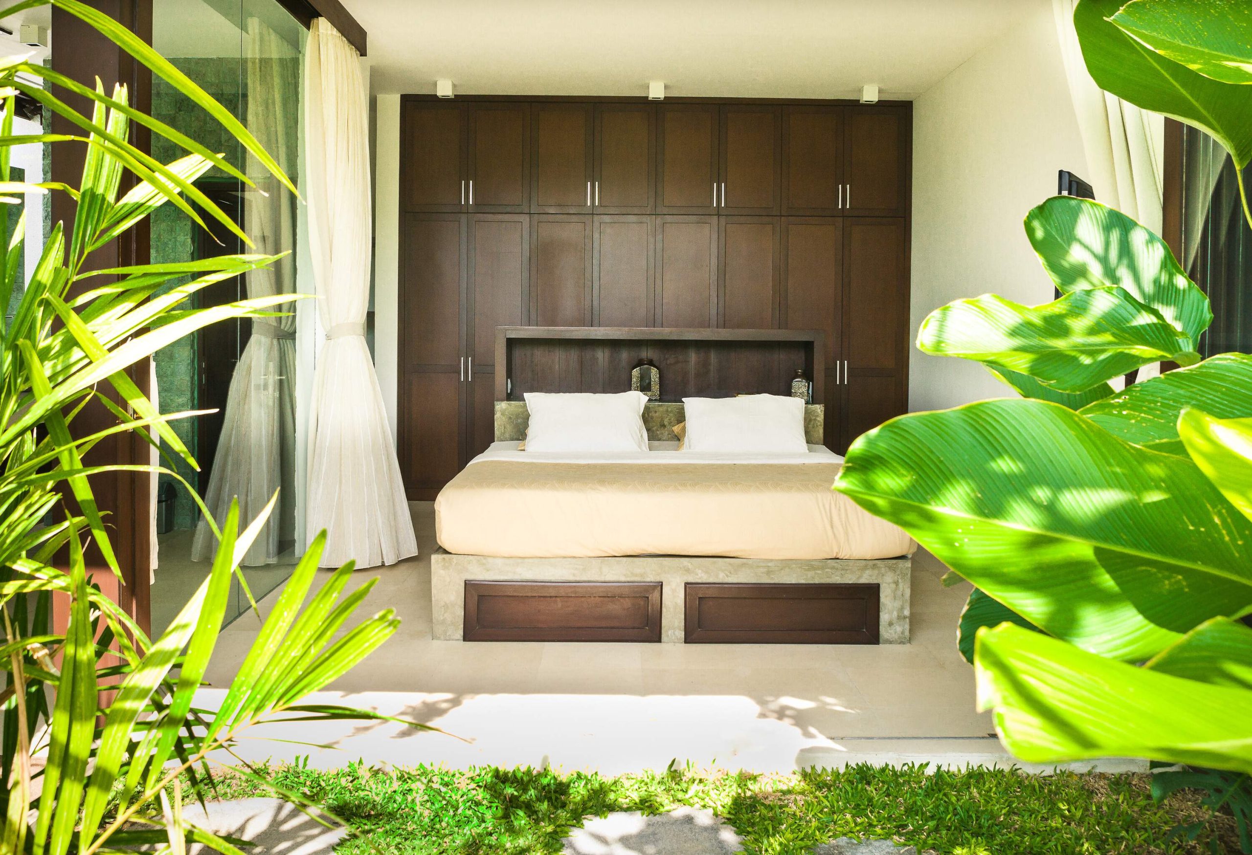 A bed in a room with open doors that leads to a garden with lush plants.