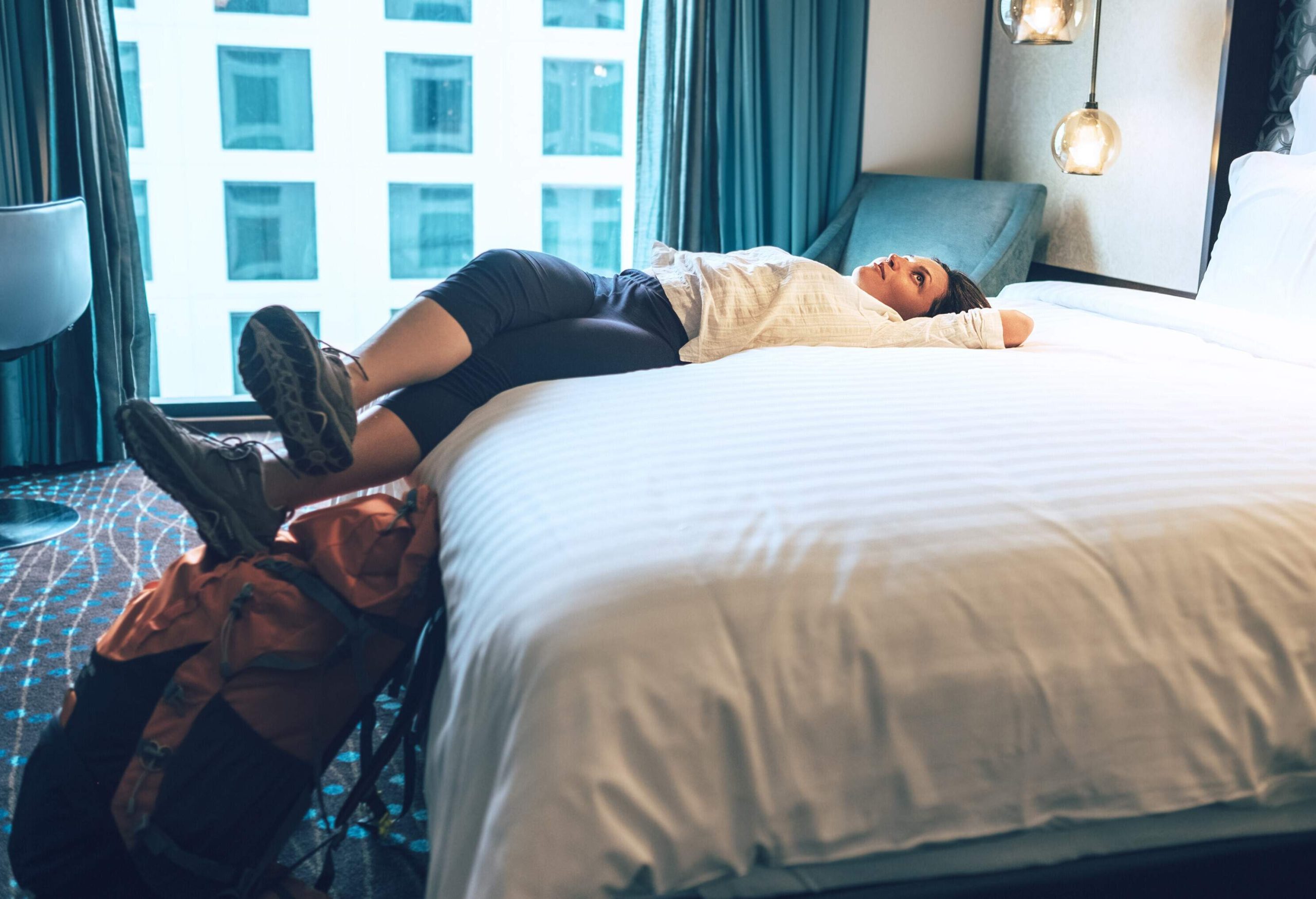 A lady backpacker lies comfortably on the white bed of her hotel room.